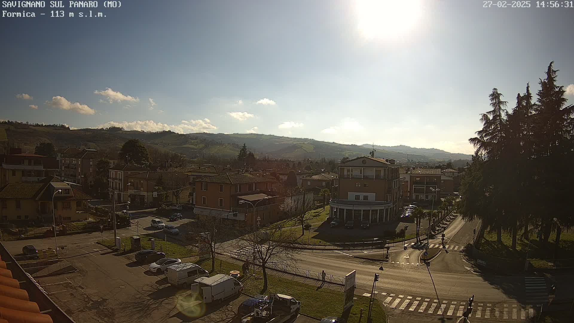 A sunny day overlooks a town nestled in a valley, with cars parked in a lot and people walking on a crosswalk.