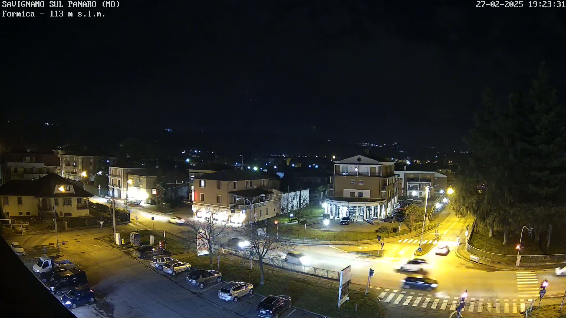 A nighttime, overhead view of a town square with cars driving through a roundabout, under a clear, dark sky.