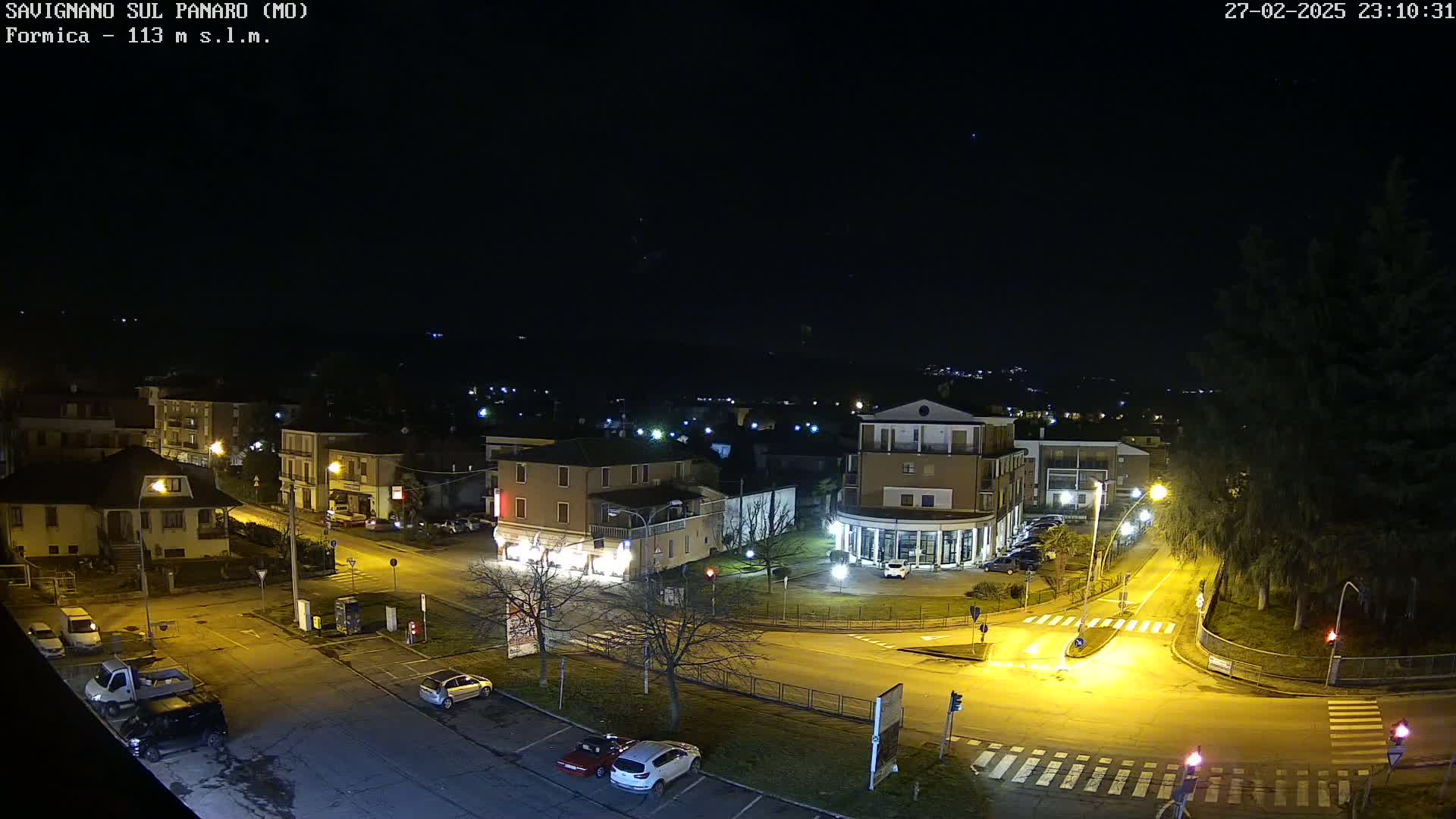 A nighttime aerial view of a town square with sparsely parked cars, illuminated by streetlights under a clear, dark sky.