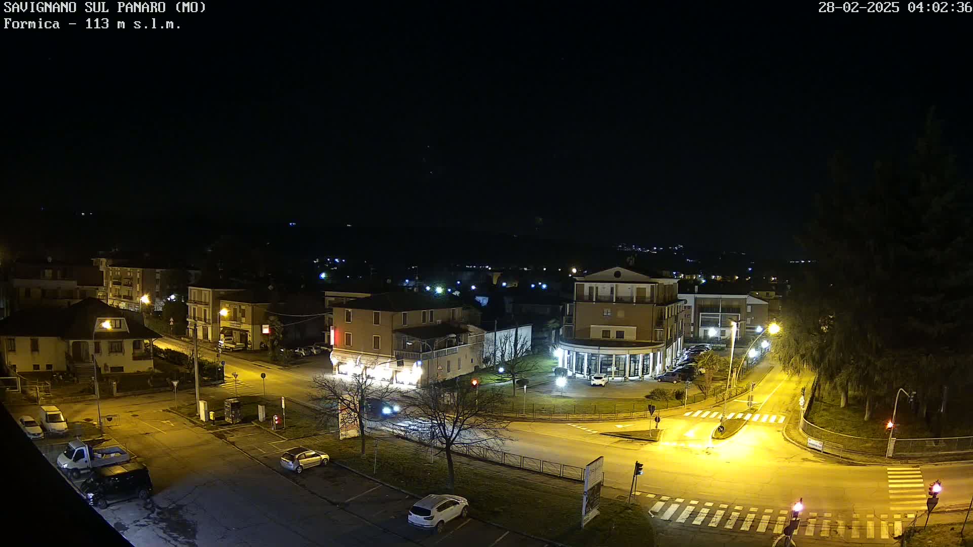 A nighttime aerial view of a town square with several buildings, cars, and streetlights, under a clear, dark sky.