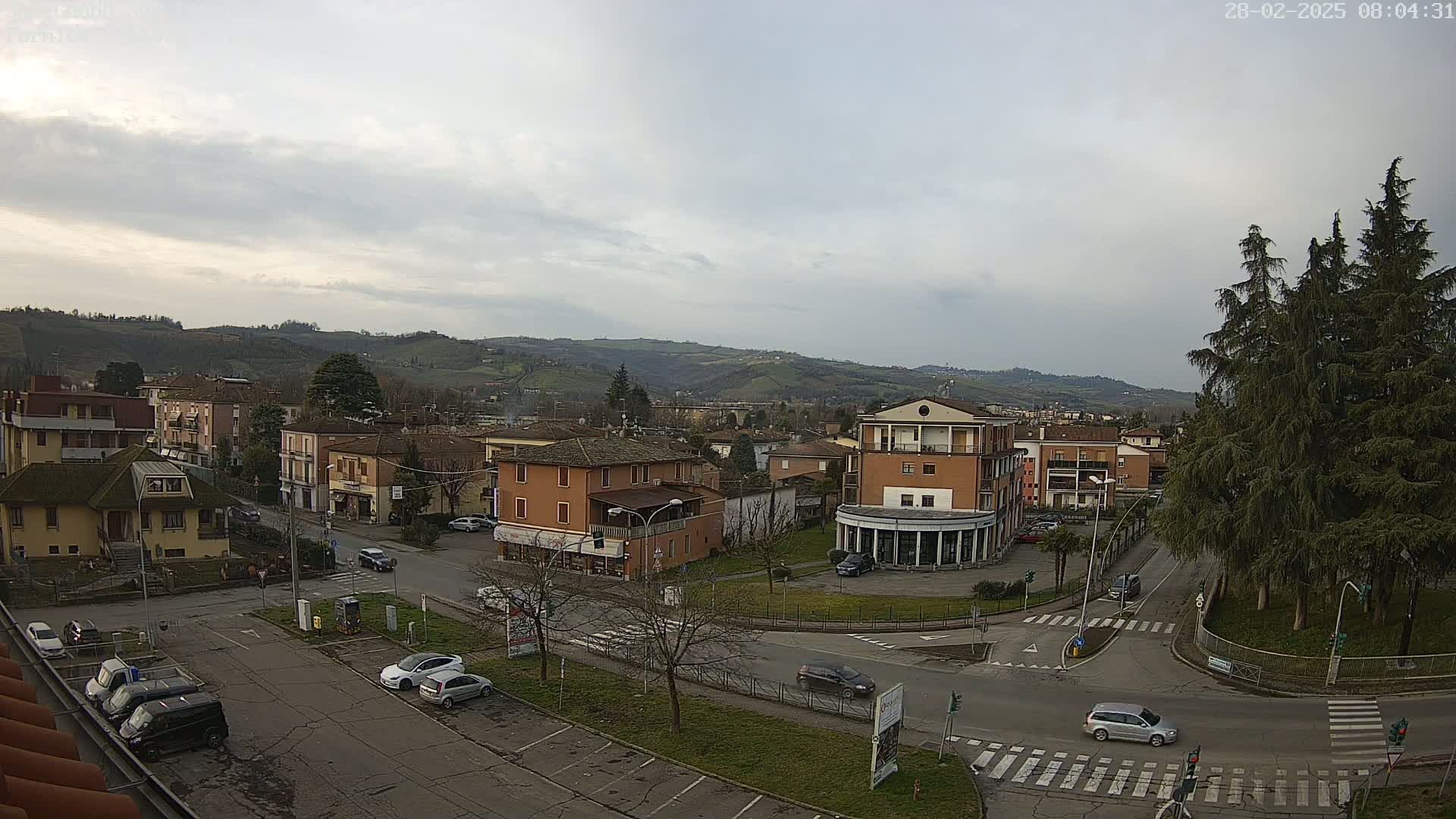A high-angle, long shot of a town square with buildings, cars, and trees under an overcast sky, with hills in the background.