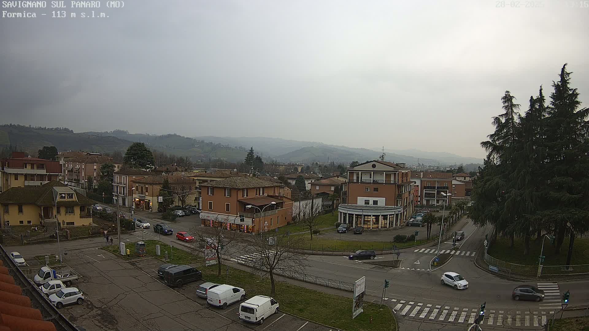 A high-angle view of a town square with cars parked and driving on a cloudy day, nestled among buildings and hills in the background.