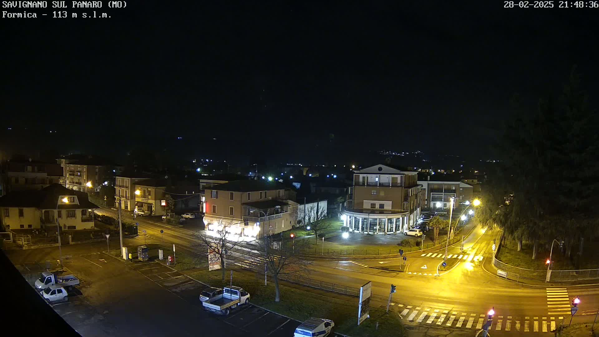 A nighttime, high-angle view of a town intersection with several buildings and parked cars, illuminated by streetlights under a clear, dark sky.