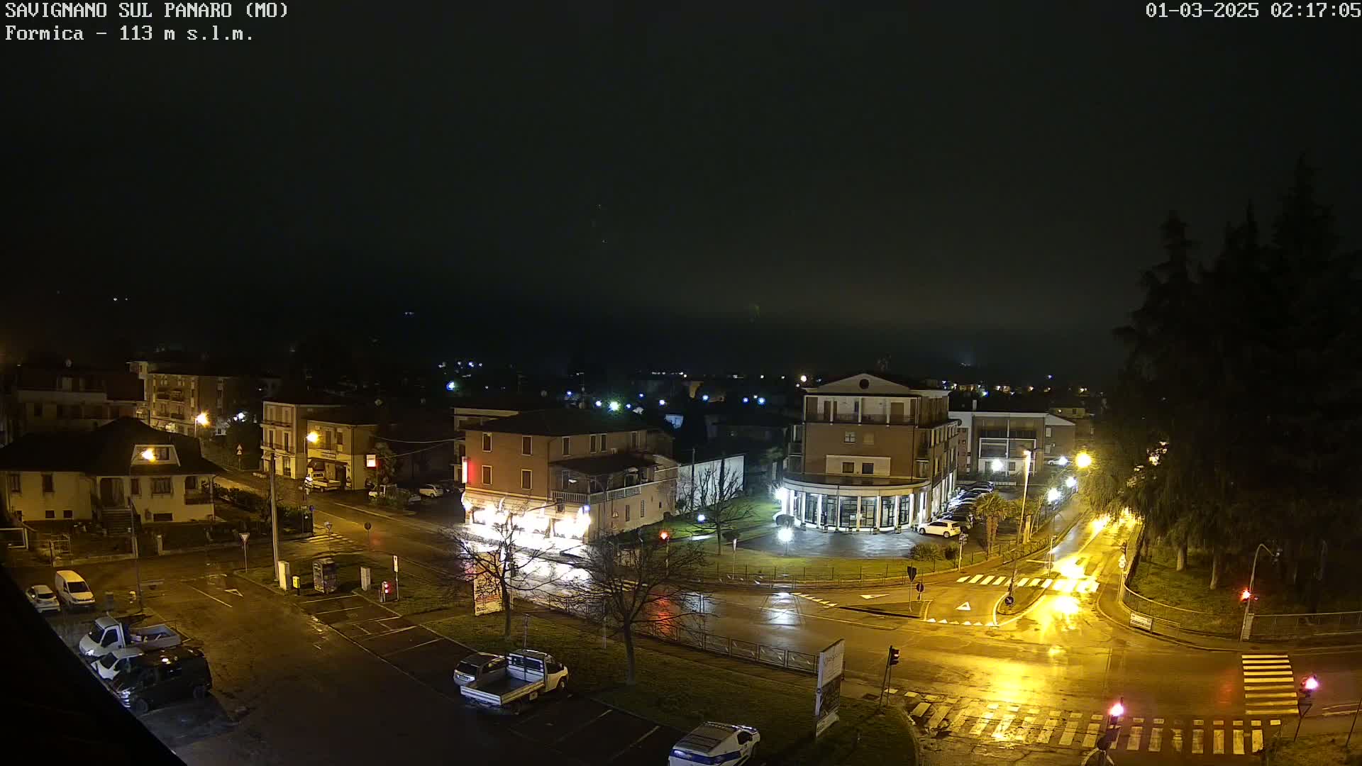 A nighttime view of a town square with wet streets and buildings illuminated under a dark, foggy sky.