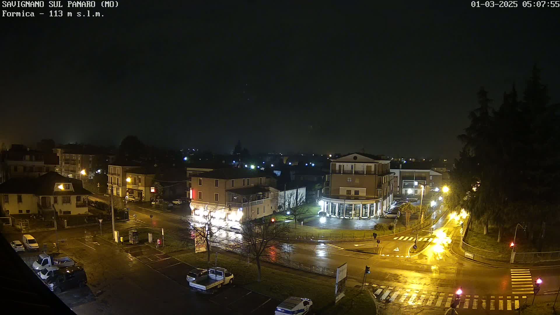 A wet, nighttime street scene shows a town square with several buildings, vehicles, and streetlights reflecting in the puddles on the roadways.