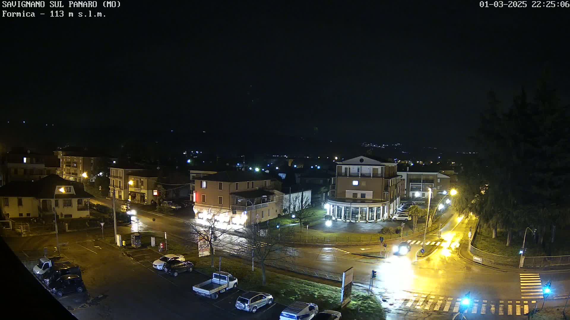 A wet, nighttime aerial view of a town square with several buildings, cars, and streetlights illuminated.