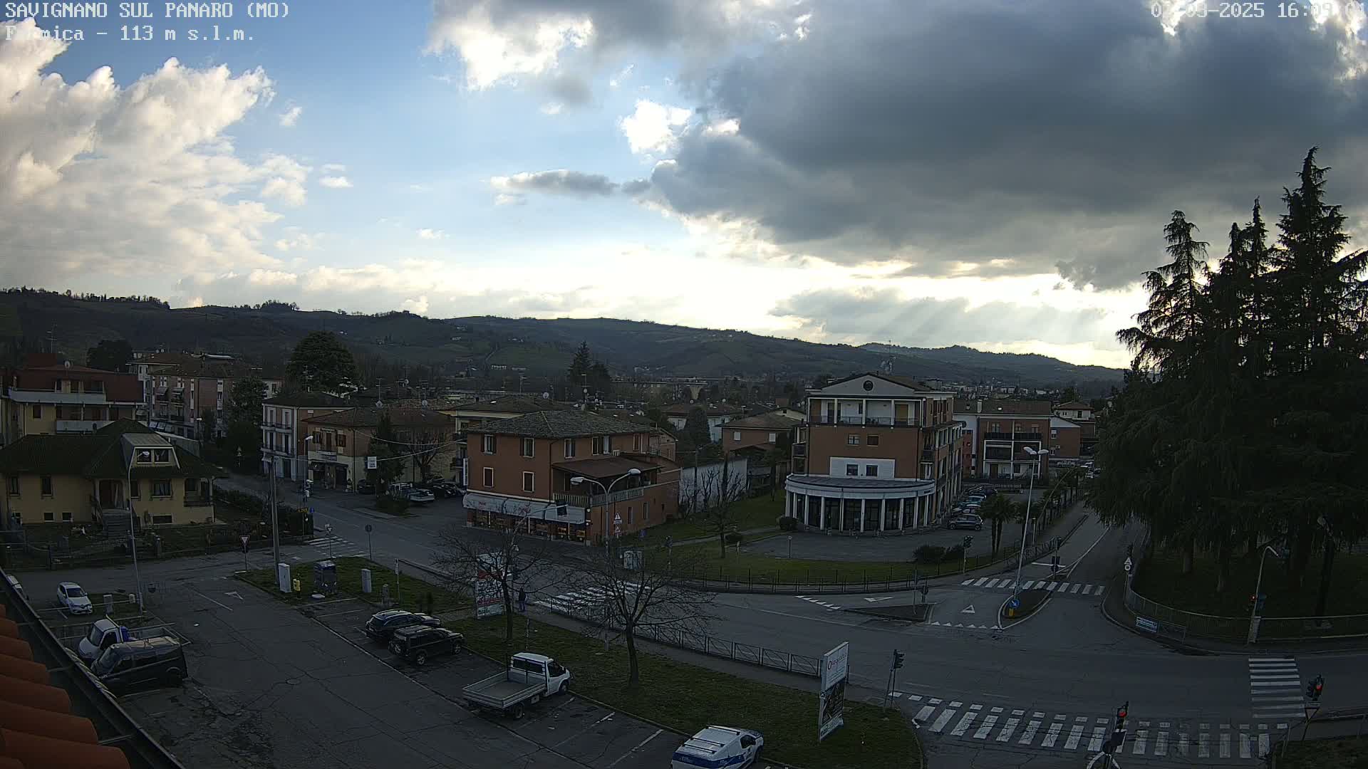 A partly cloudy day reveals a view of a town nestled in a valley, with cars parked along streets and roads intersecting at a roundabout.