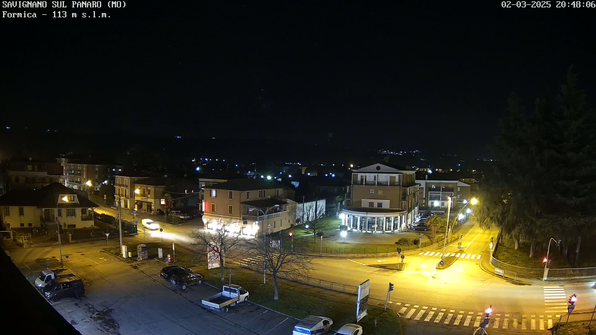 A nighttime, overhead view of a town square with buildings, vehicles, and streetlights illuminating the scene under a clear, dark sky.