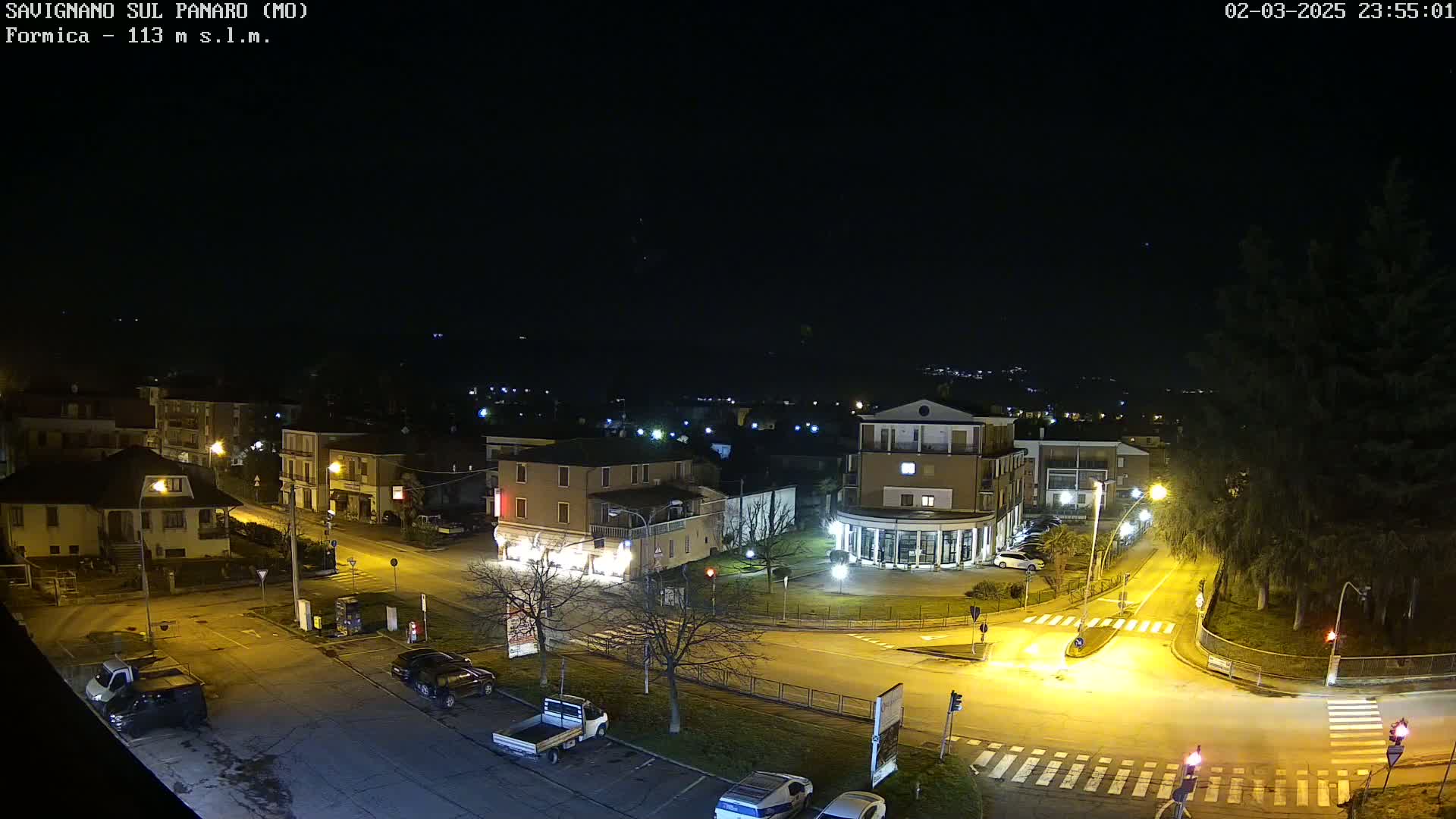 A nighttime aerial view of a town square with buildings, parked cars, and streetlights illuminating the intersection under a clear, dark sky.