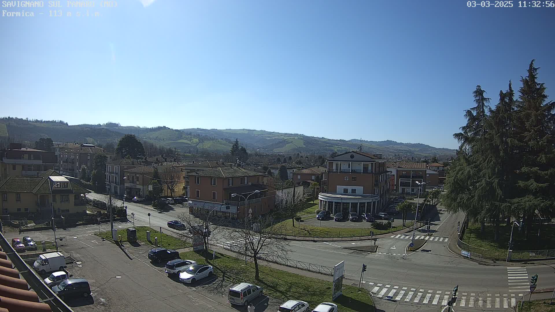 A sunny day overlooks a town with several buildings and cars parked along the streets, rolling hills in the background.