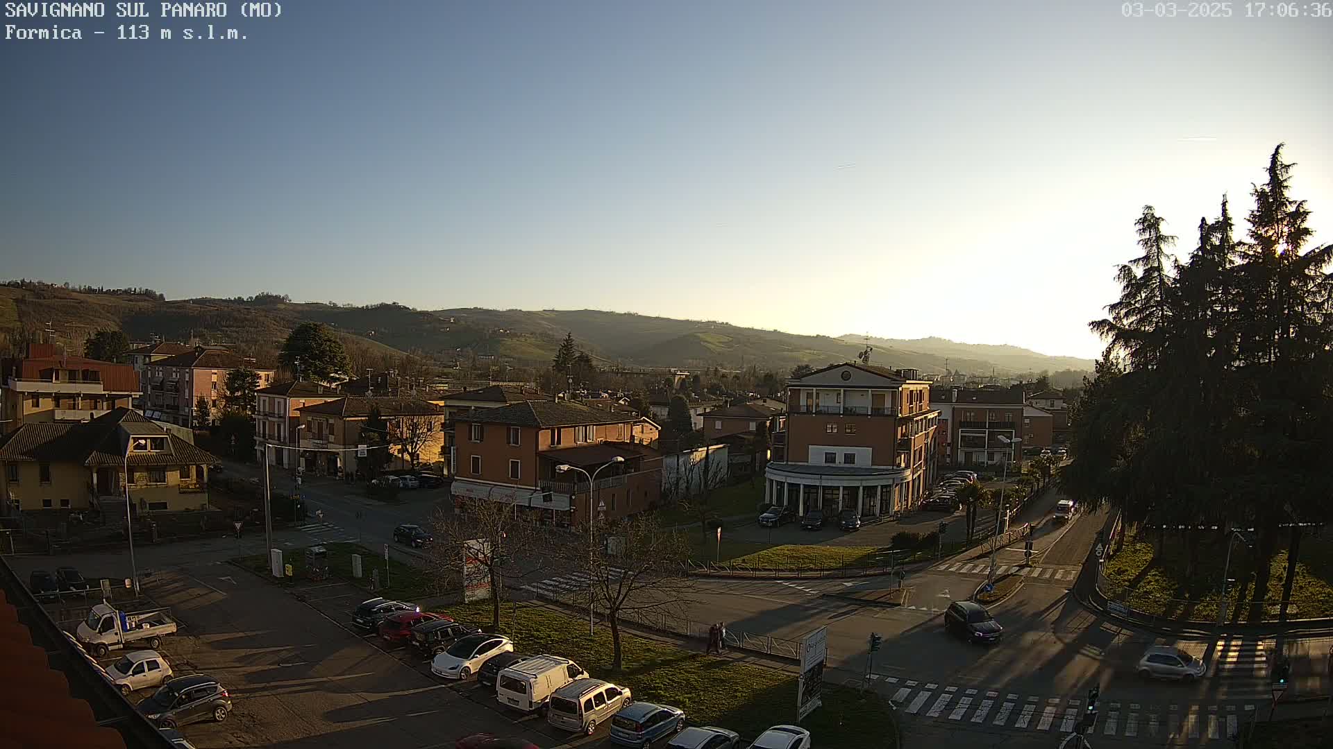 A high-angle, long shot of a town nestled in a valley, bathed in the warm light of a setting sun under a clear sky, with cars parked in a lot and driving on nearby streets.