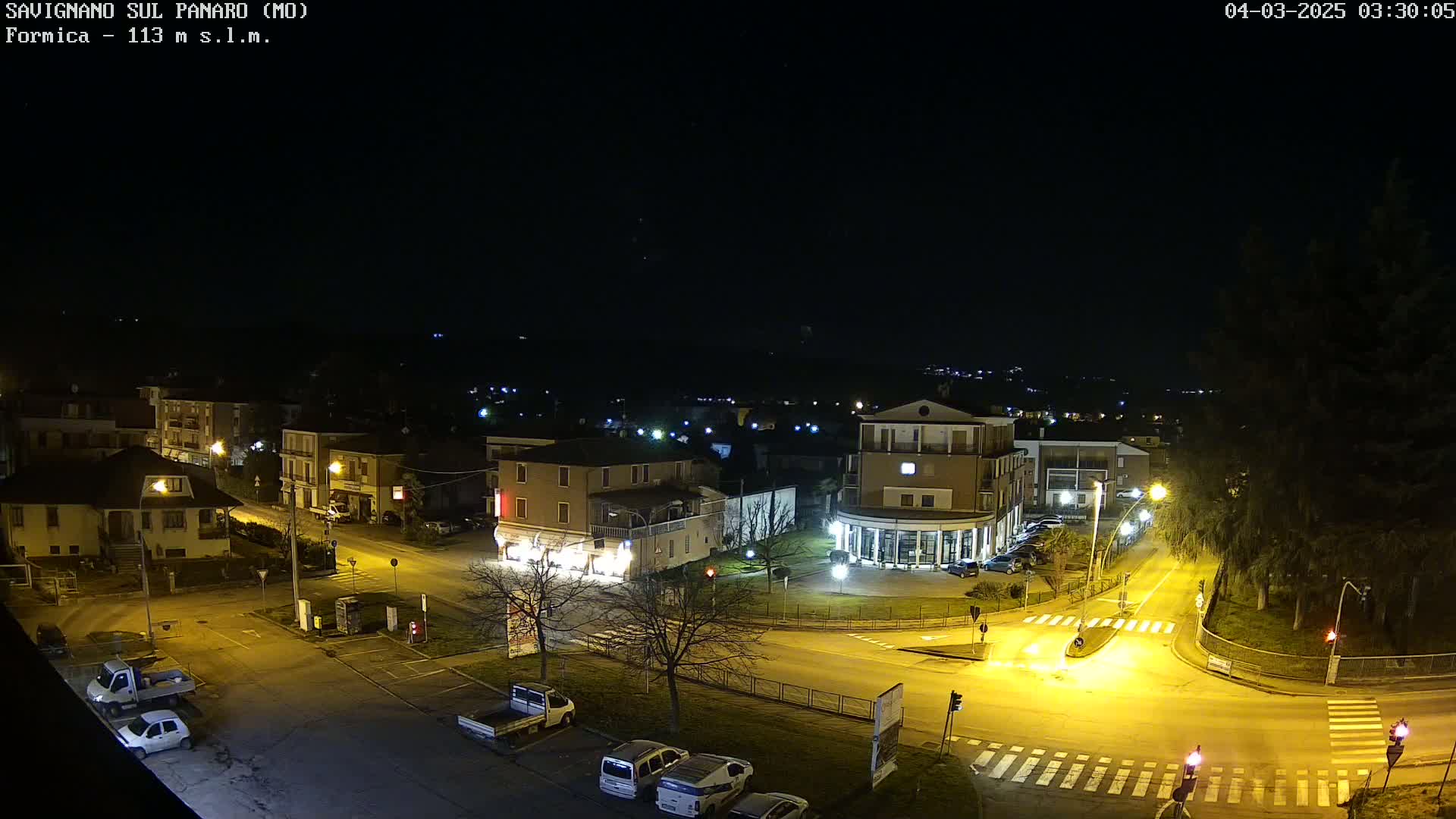 A nighttime, overhead view of a town square with several buildings, parked cars, and streetlights illuminating the area under a clear, dark sky.