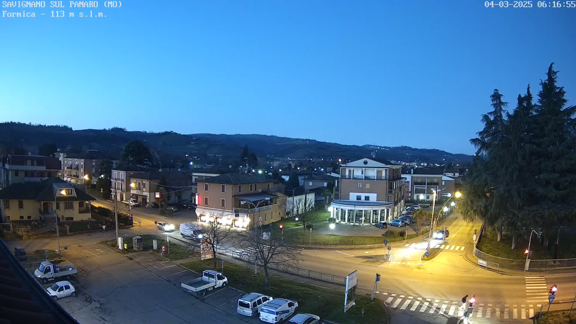 A nighttime view of a town square with buildings, cars, and streetlights, under a twilight sky.