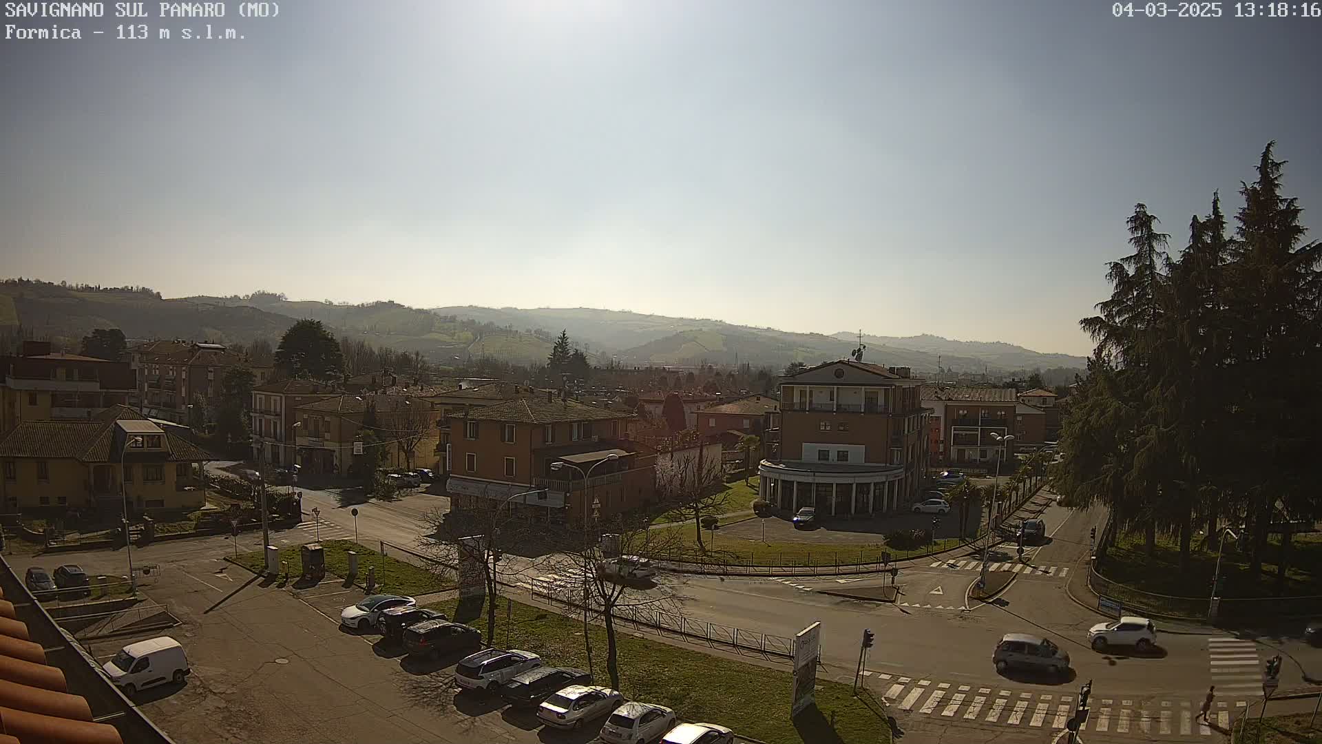 A sunny view of a town nestled in a valley, with cars parked along the streets and hills visible in the background.