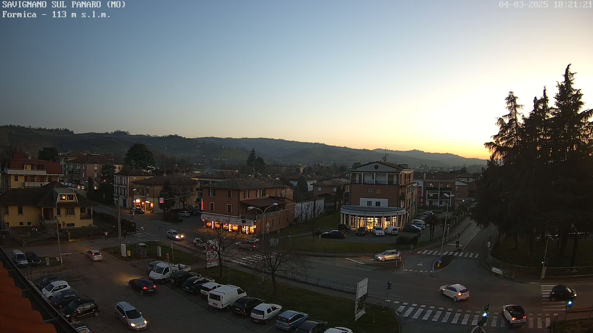 A twilight view of a town square with cars parked and driving, nestled among buildings at the foot of hills under a clear, dusky sky.