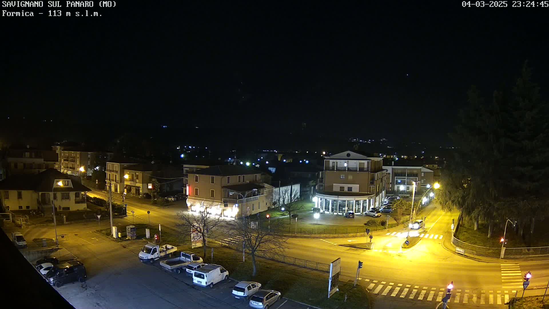 A nighttime aerial view of a town square with several buildings, vehicles parked along the streets, and streetlights illuminating the scene under a clear, dark sky.