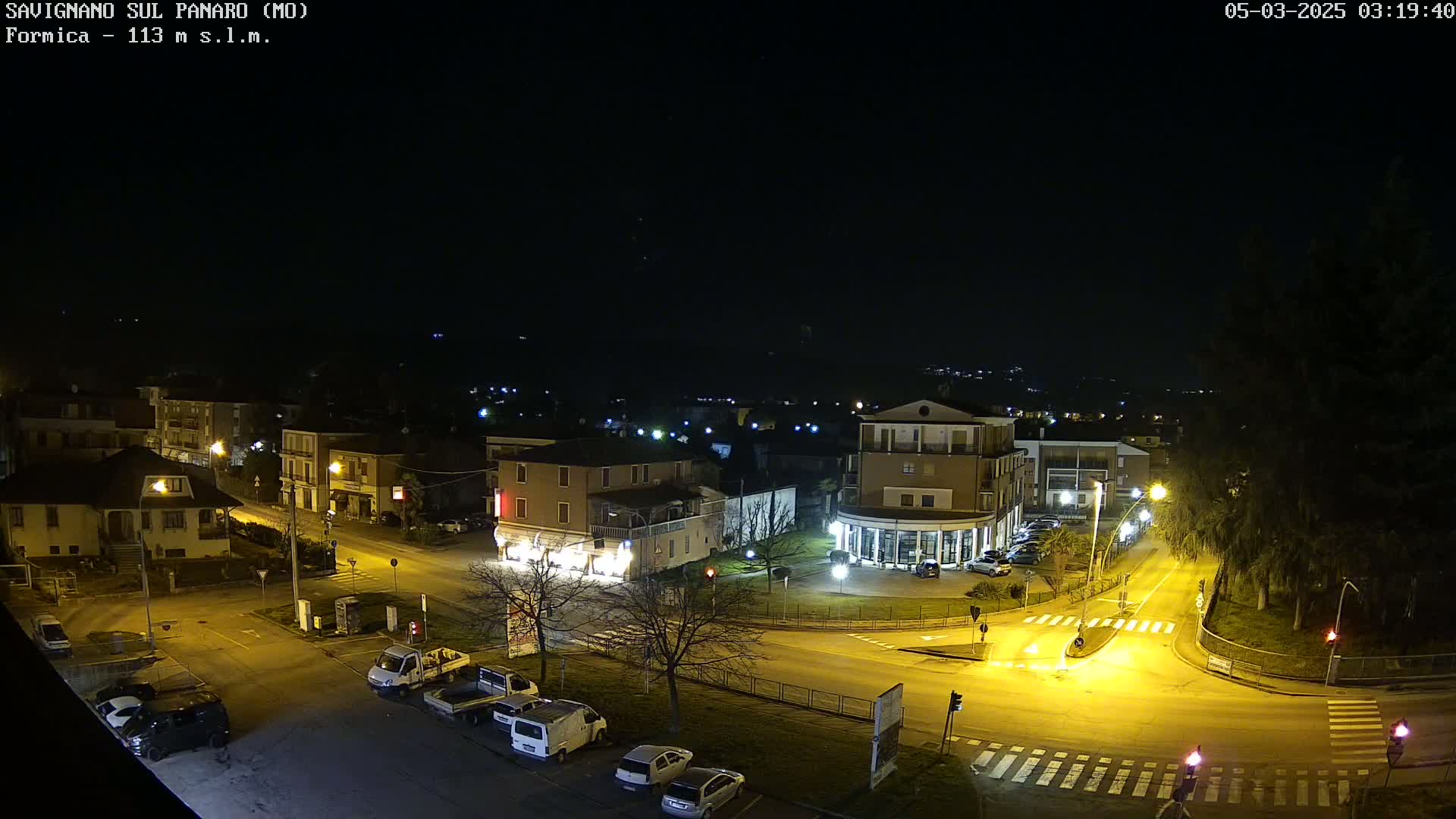 A nighttime aerial view of a town square with parked cars and illuminated buildings under a clear, dark sky.