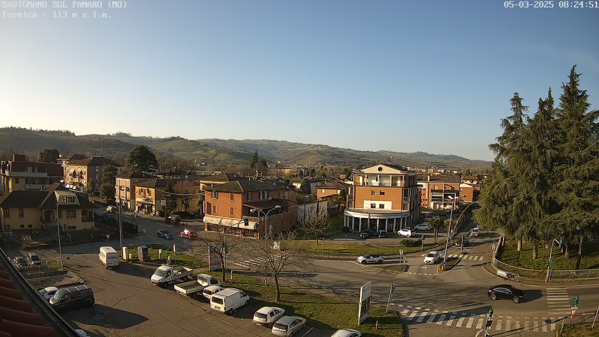A sunny day reveals a town square in a hilly region, with several buildings, cars, and a large evergreen tree visible.