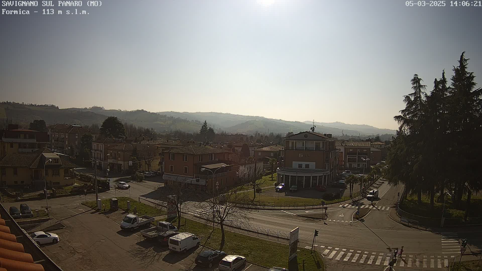 A sunny day reveals a view of a town nestled in a valley, with roads and buildings visible, including a few parked cars and some tall evergreen trees.