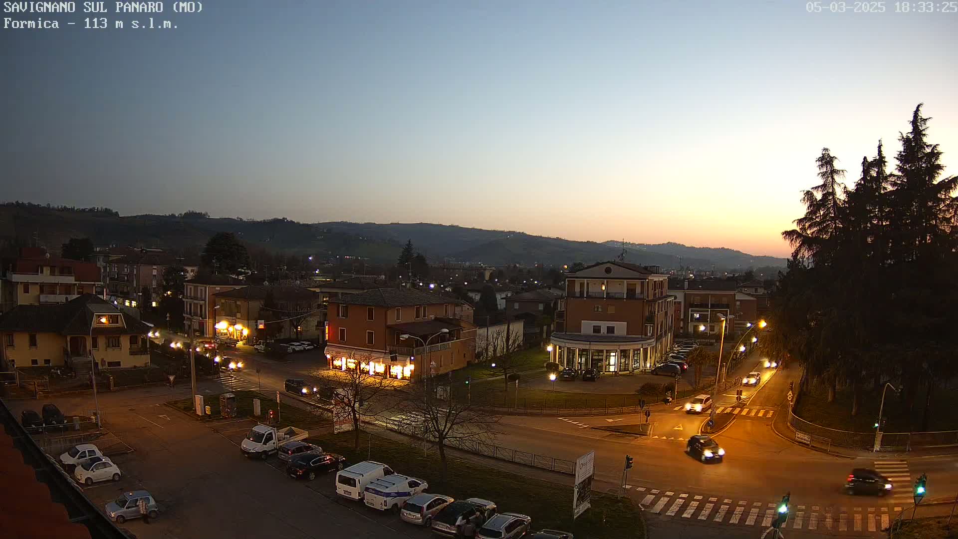 A twilight view of a town square with cars and buildings illuminated by streetlights, under a clear, dusky sky.
