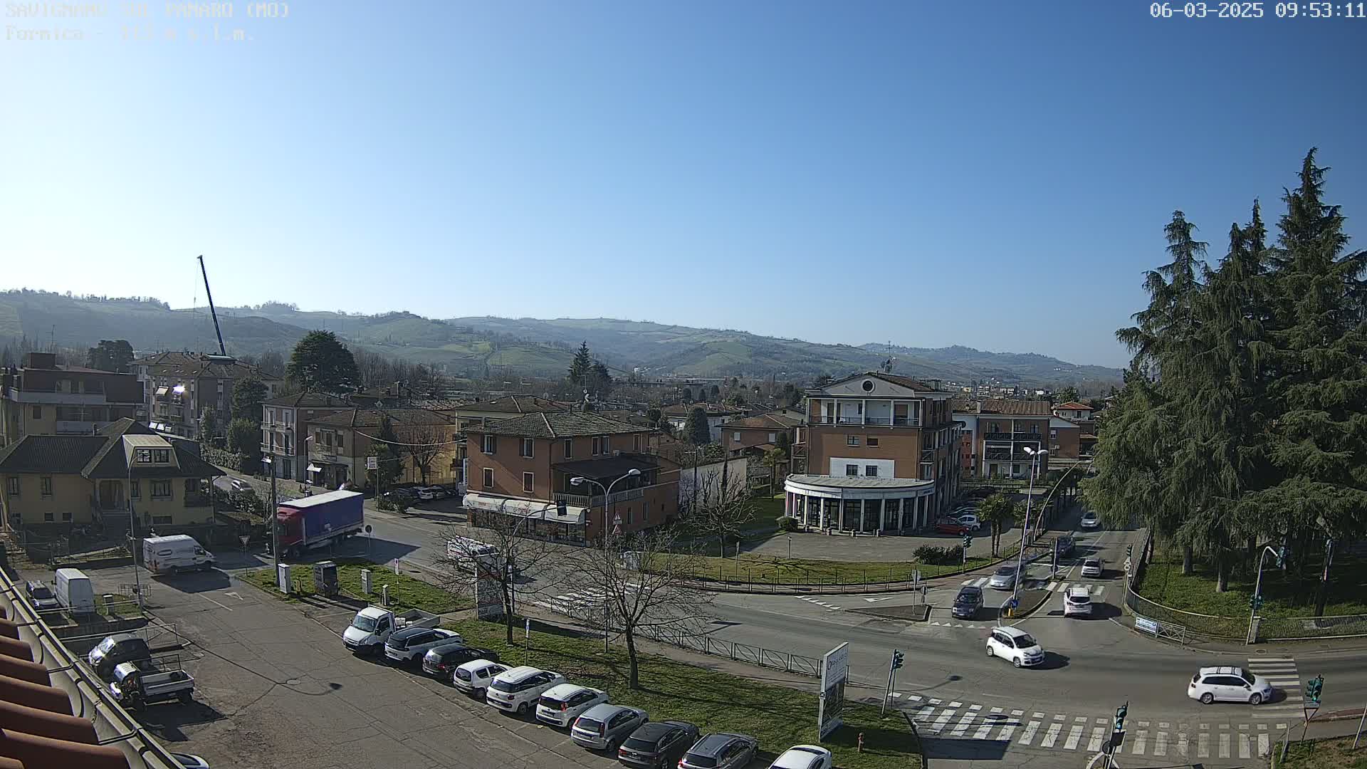 A sunny day reveals a town nestled in a valley, with cars traversing a roundabout surrounded by buildings and evergreens, under a clear blue sky.