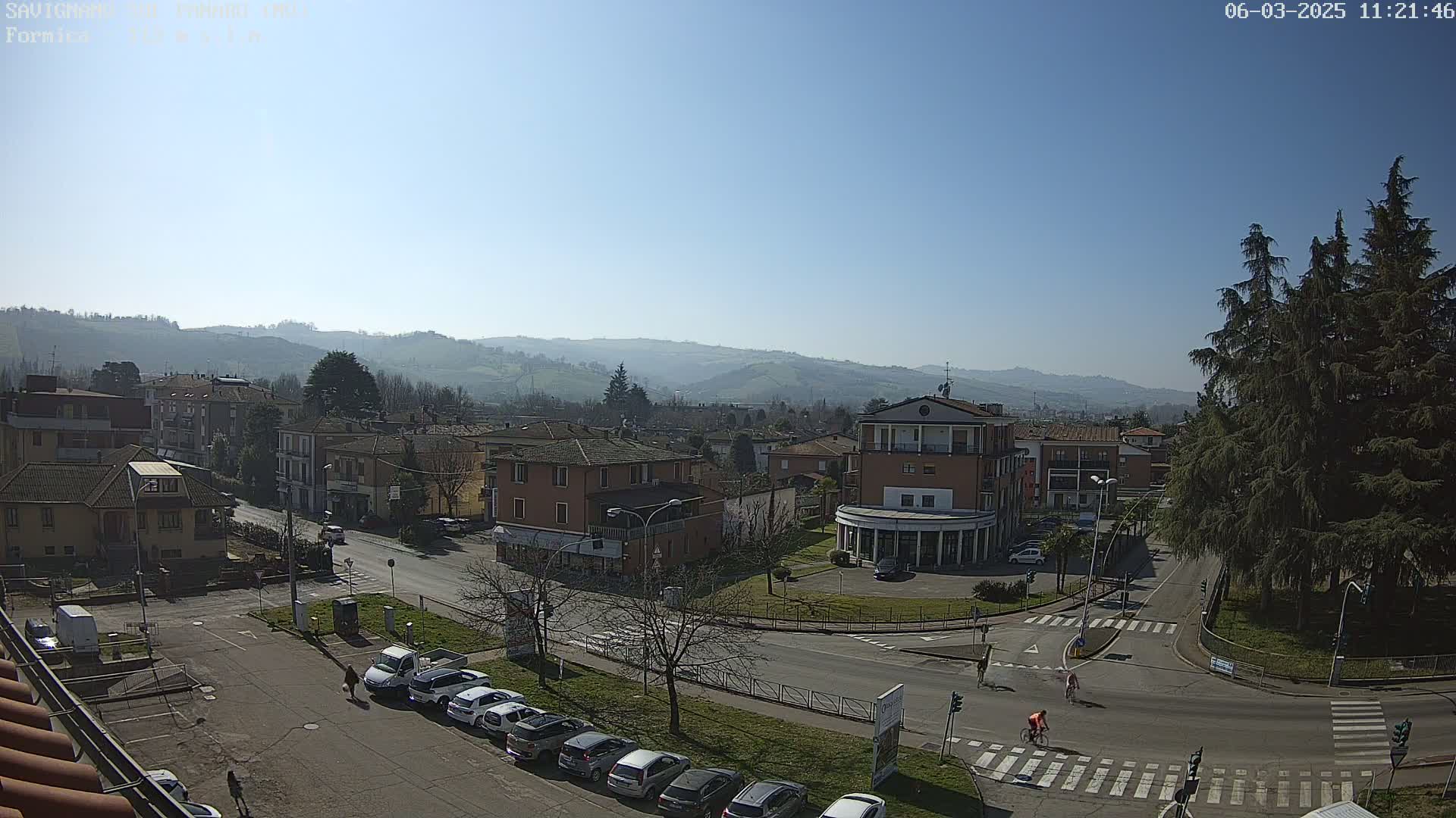 A sunny day reveals a town's streetscape nestled among rolling hills, with cars parked along the roadside and a few pedestrians and cyclists visible.