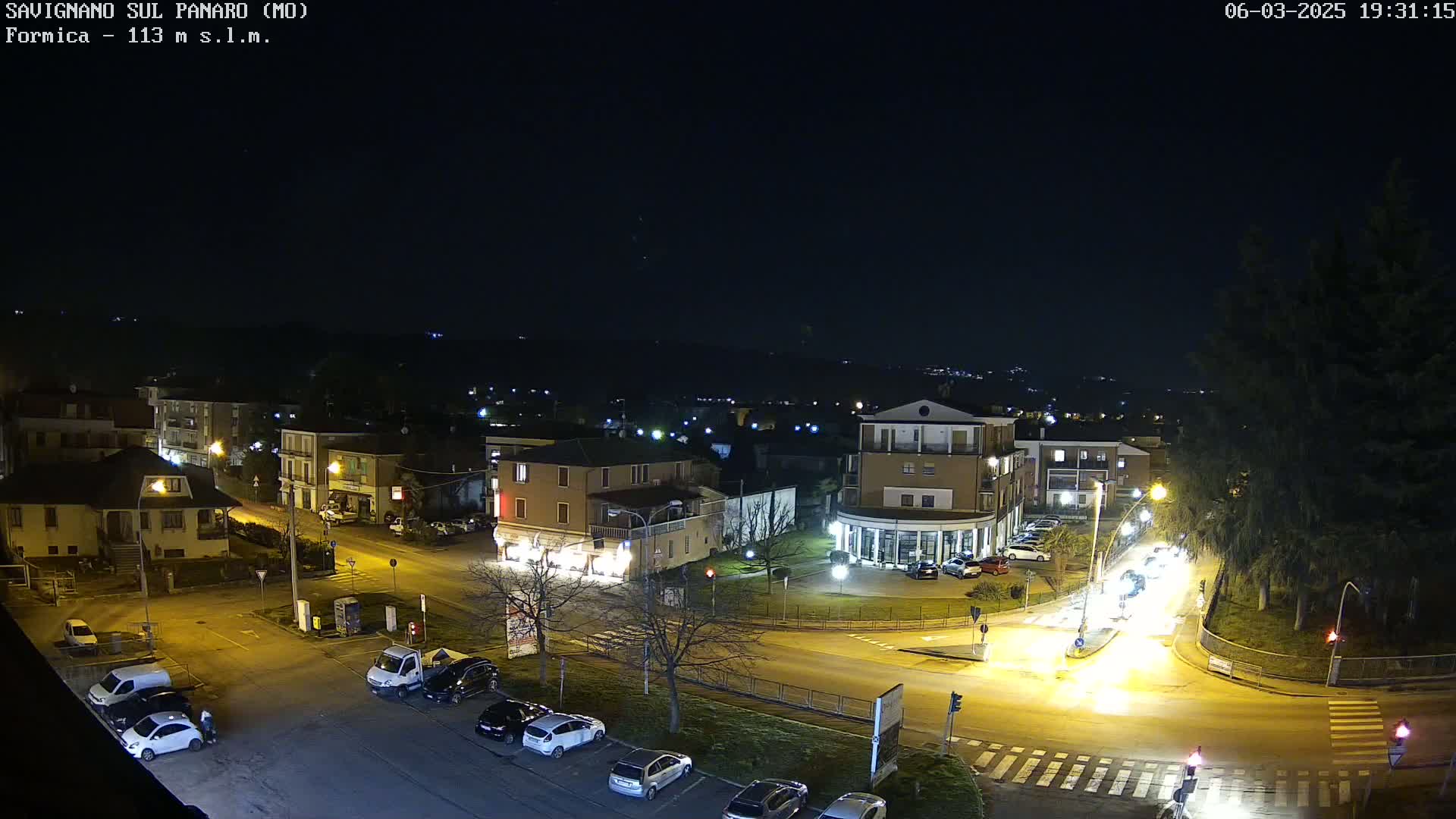 A nighttime, high-angle view of a town square with cars parked and driving around a roundabout, under a clear, dark sky.