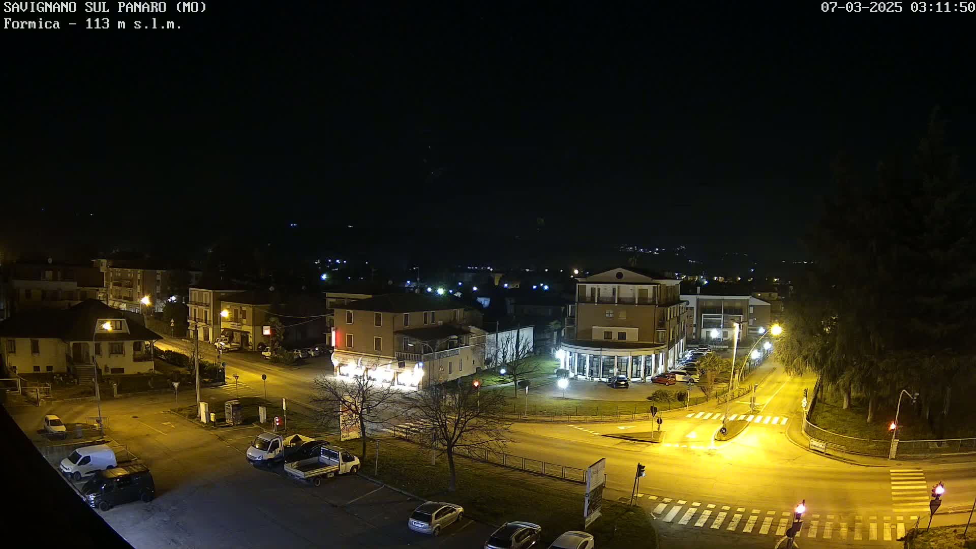 A nighttime, overhead view of a town square with illuminated streets, buildings, and parked cars under a clear, dark sky.