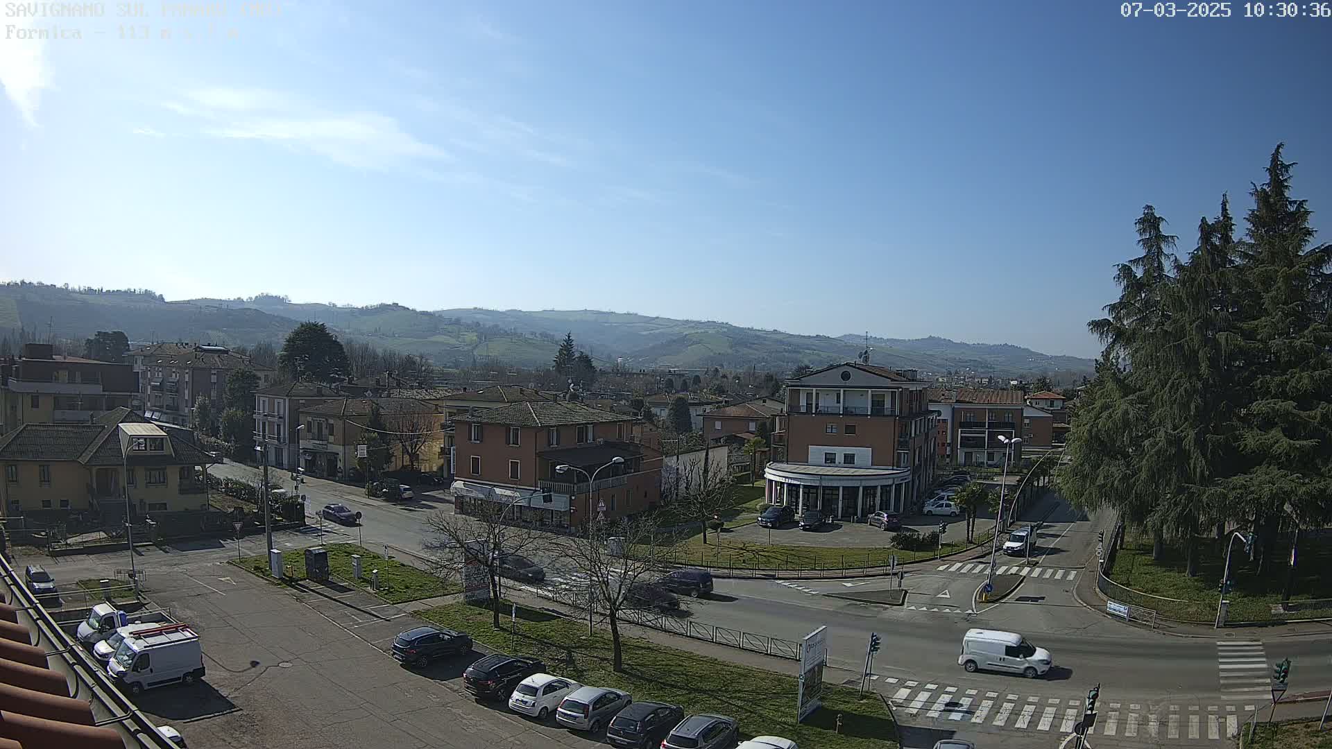 A sunny day reveals a town nestled in a valley, with cars parked along streets and a few vehicles traversing a crossroads near evergreen trees, all under a mostly clear sky.