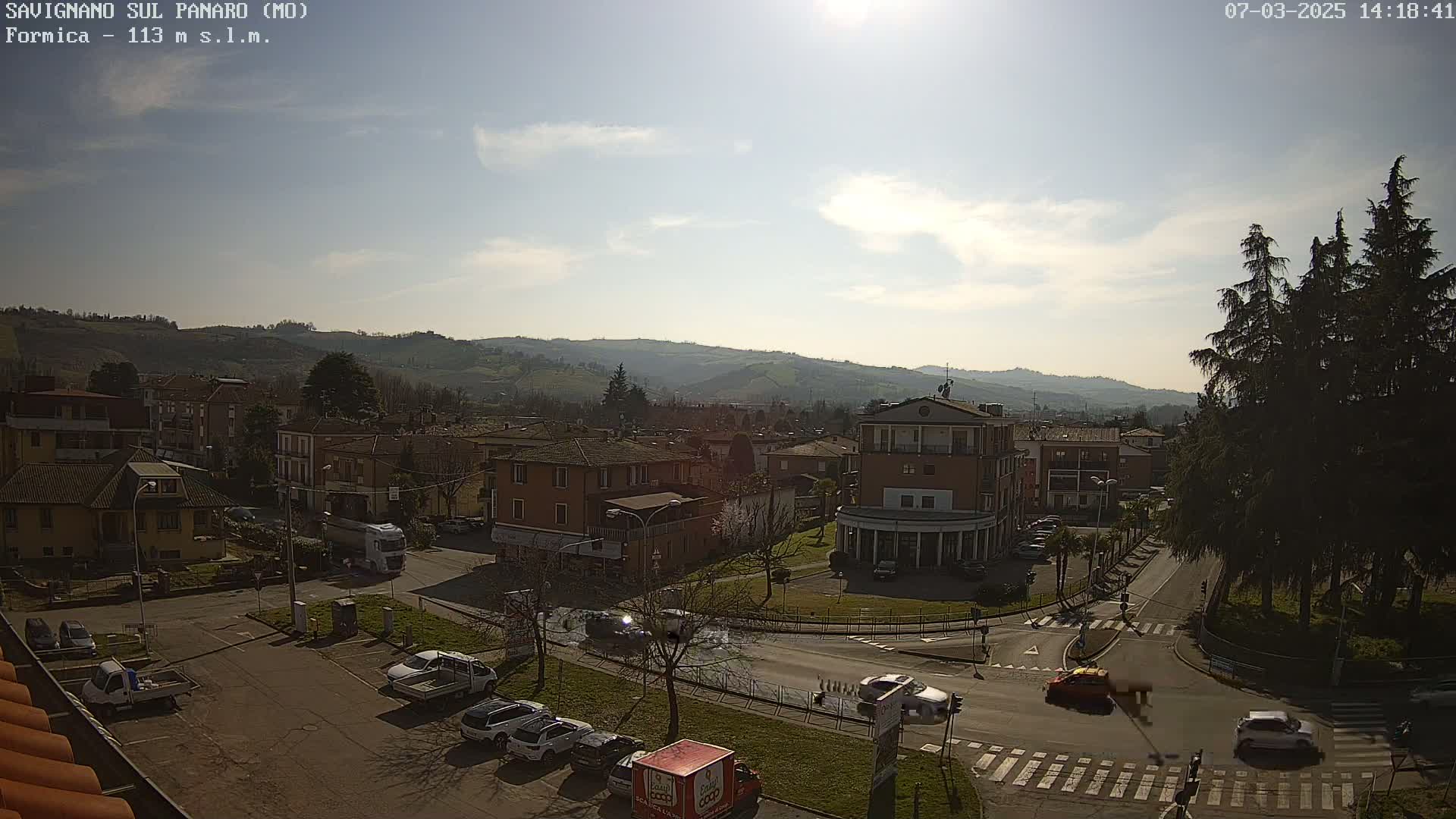 A sunny day overlooks a town nestled in a valley, with cars parked and driving on roads that intersect near a roundabout.