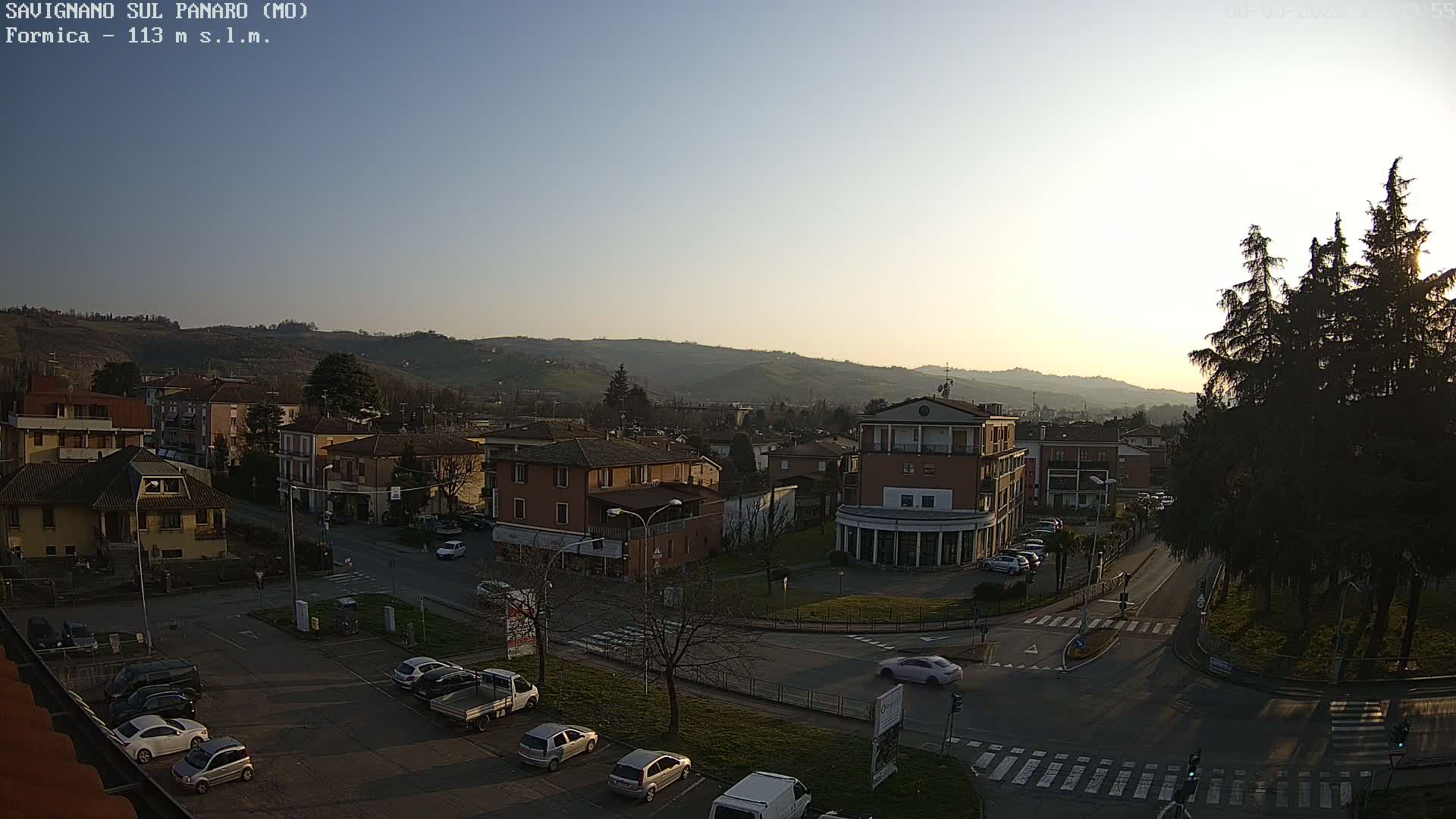 An aerial view of a town at sunset, under clear skies, showing residential buildings, roads, parked cars, and a few pedestrians.