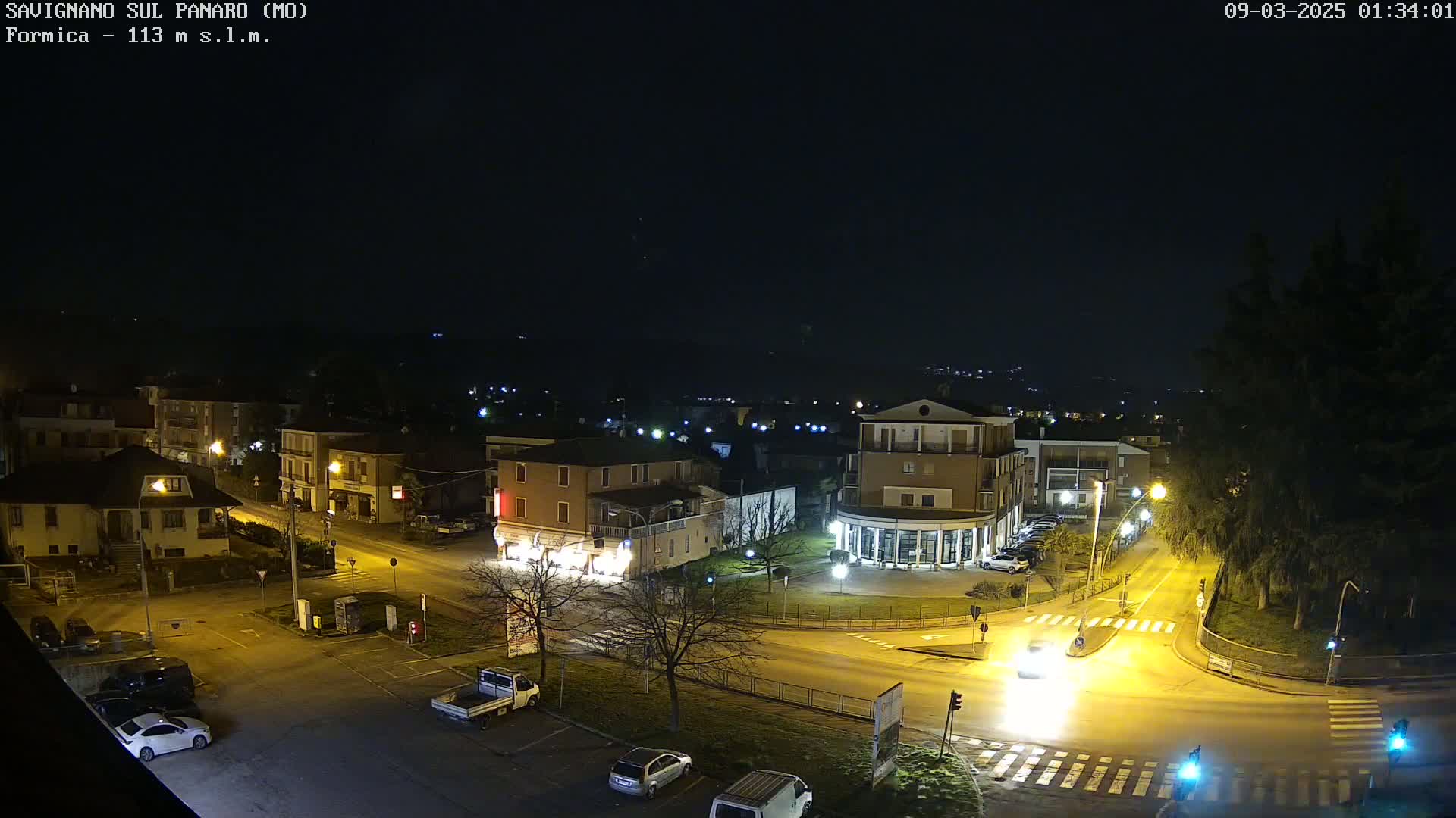 A nighttime, high-angle view of a town's intersection with several buildings and cars illuminated by streetlights.