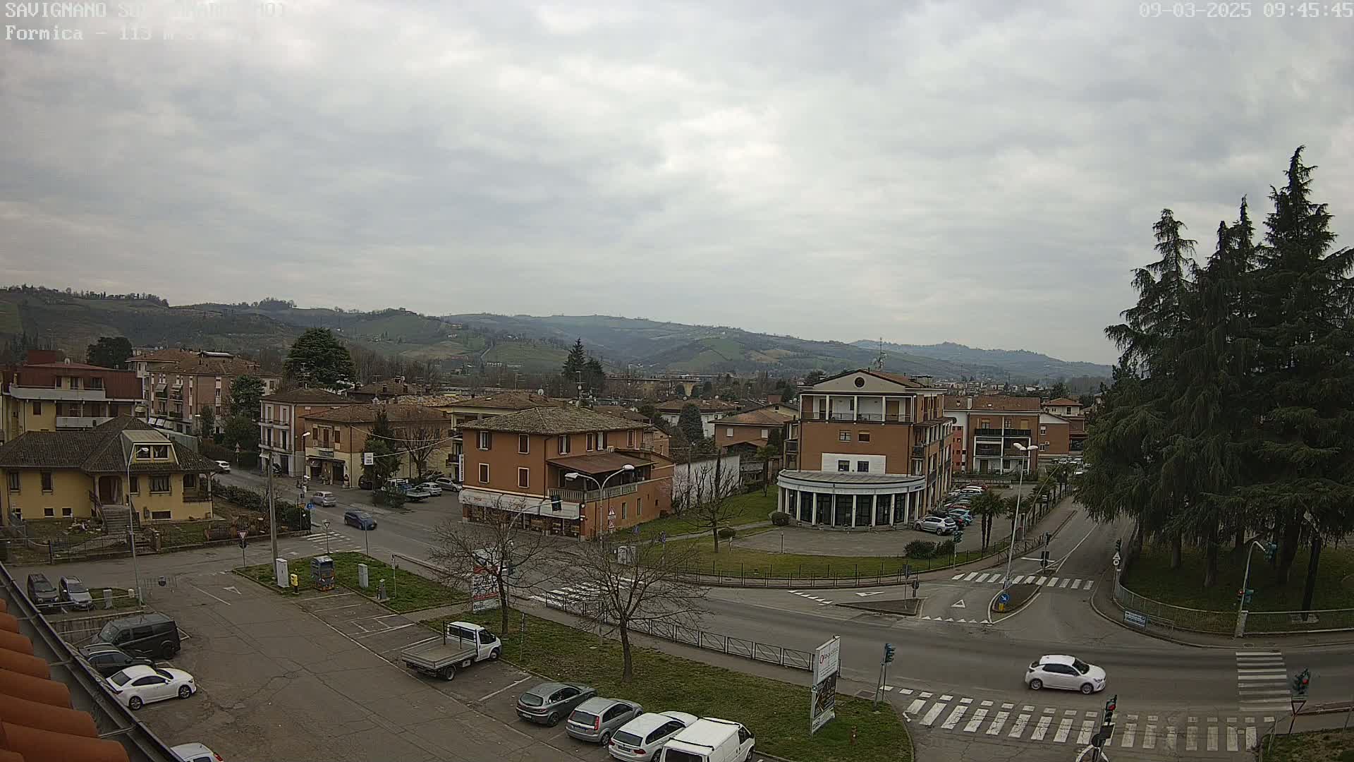 A mostly cloudy day overlooks a town square with cars parked and driving, several buildings, and a background of rolling hills.