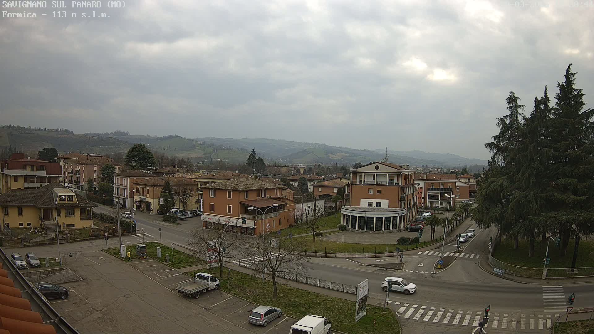 An overcast day reveals a town square with several buildings, cars, and a small park, nestled in a valley surrounded by rolling hills.