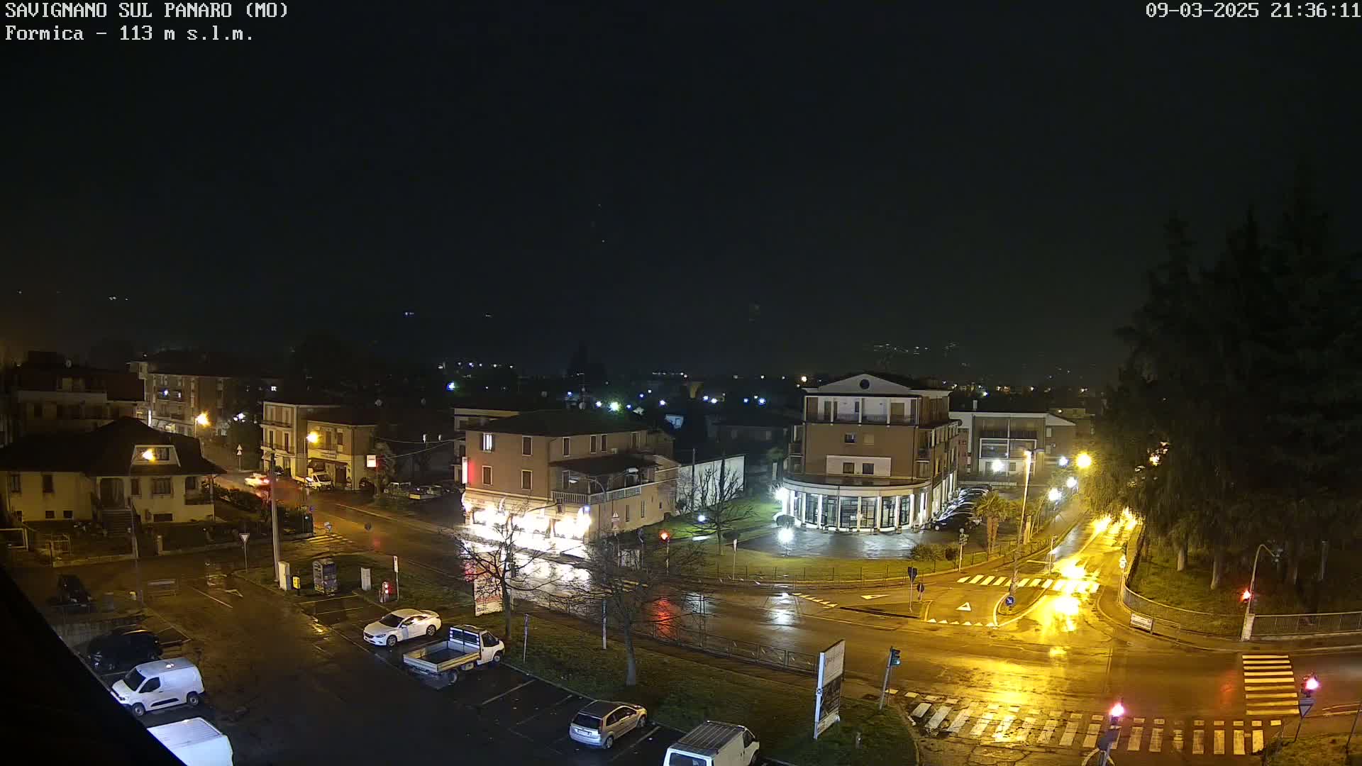 A wet, nighttime, high-angle view of a town square with buildings, vehicles, and streetlights illuminated under a dark sky.