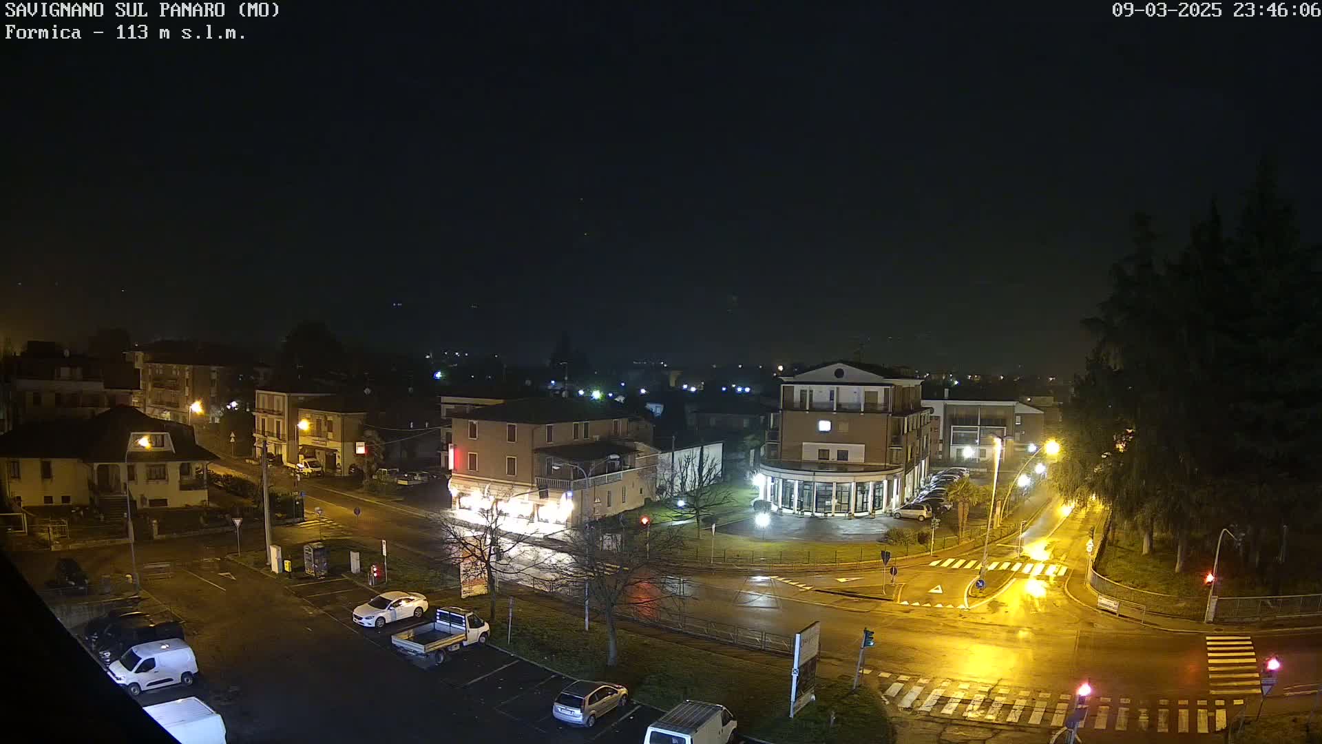 A nighttime view of a town square with wet roads and sparsely parked cars under a dark, possibly foggy sky.