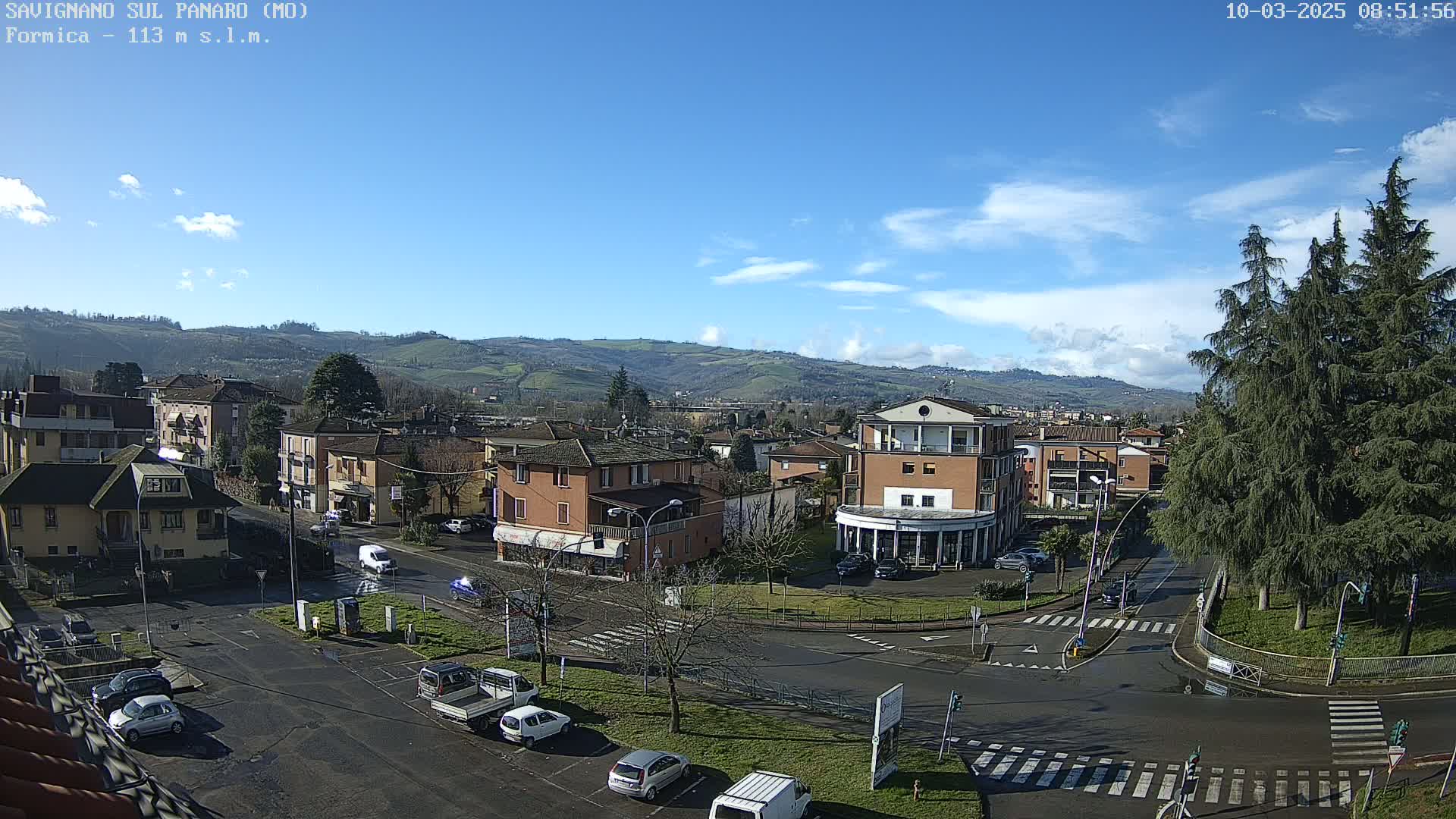A sunny day reveals a small town nestled in a valley, with cars parked in a lot and driving on roads that intersect near a roundabout, and evergreen trees in the foreground.