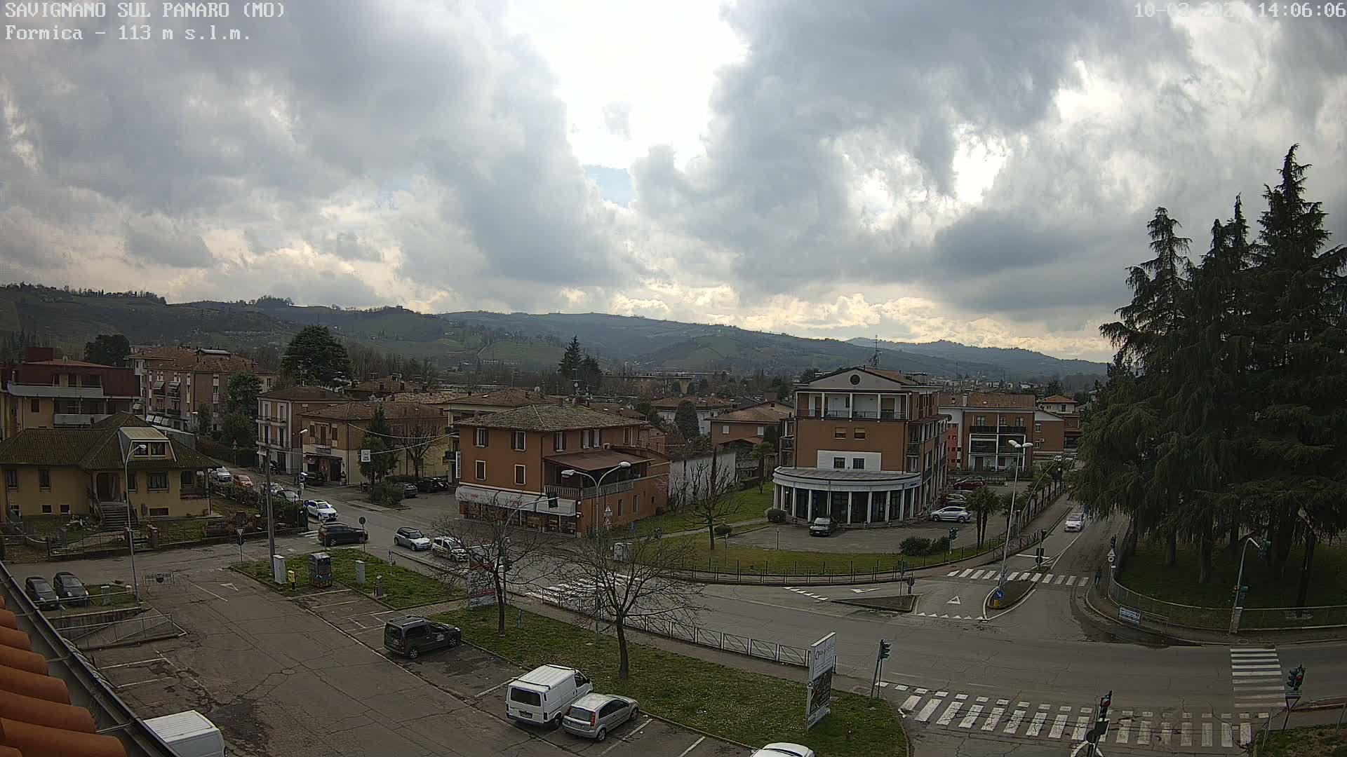 A mostly cloudy day reveals a view of a town square and surrounding buildings nestled in a valley, with mountains visible in the background.