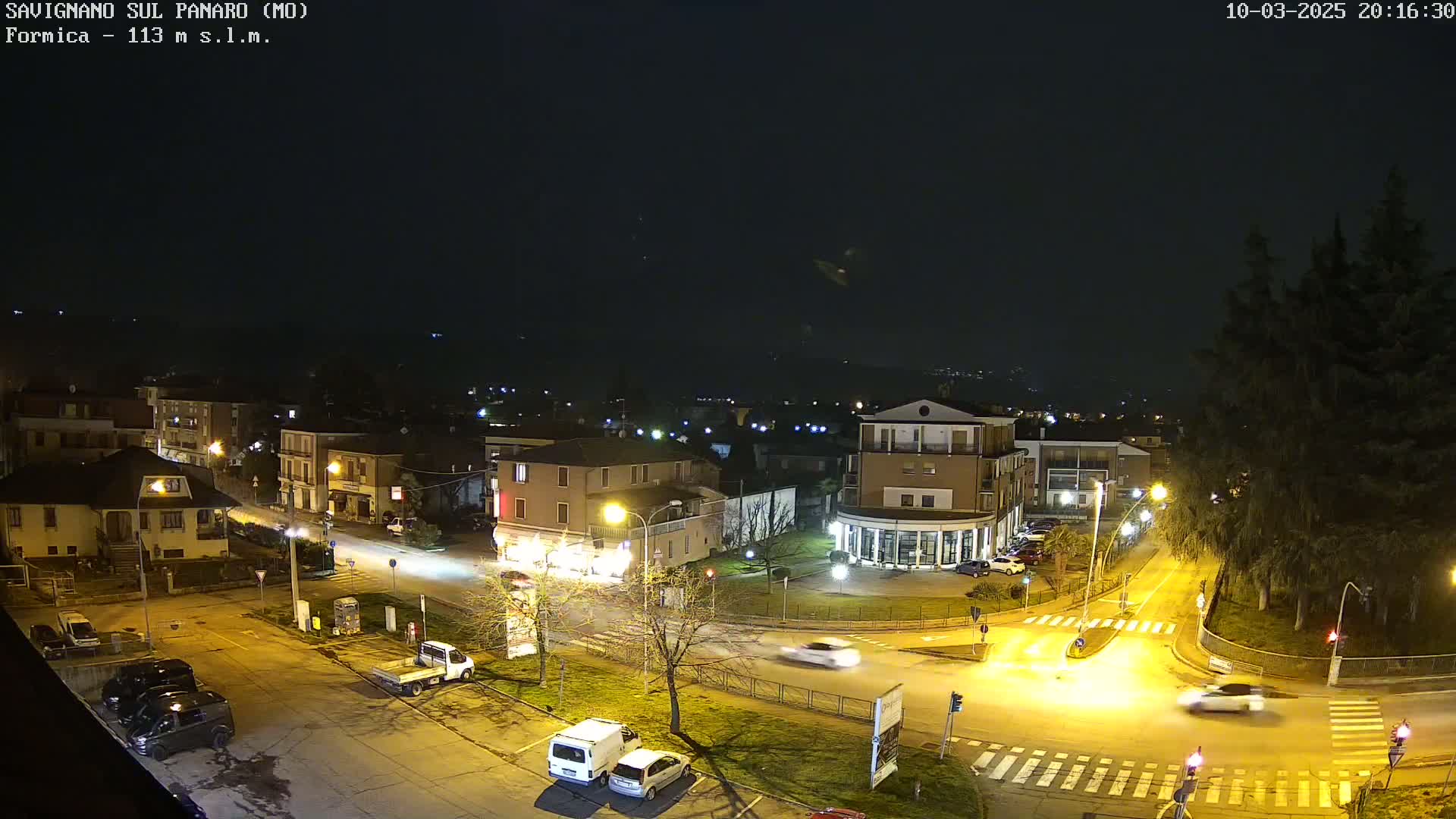 A nighttime, overhead view of a town intersection with several cars and buildings illuminated by streetlights under a clear, dark sky.