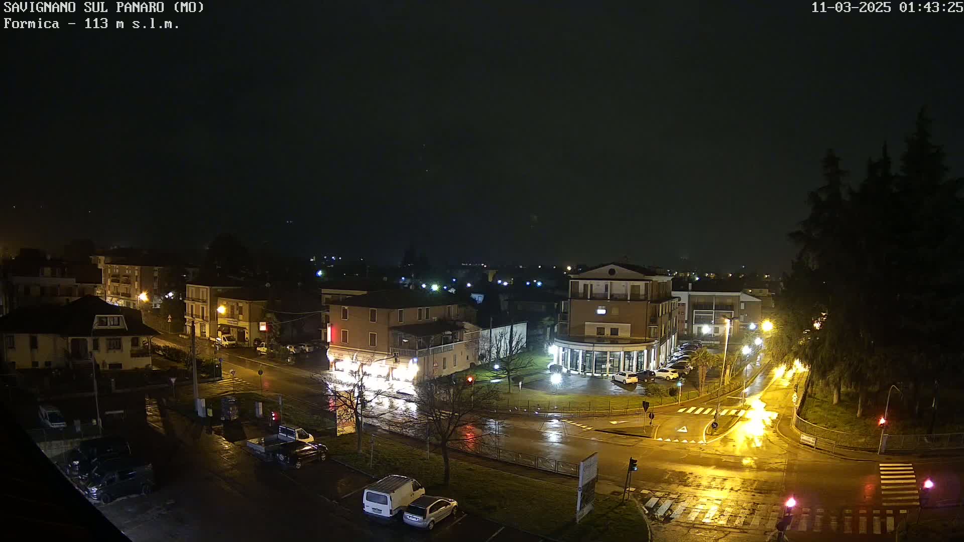 A rain-slicked town square at night, illuminated by streetlights, shows several buildings and parked cars.