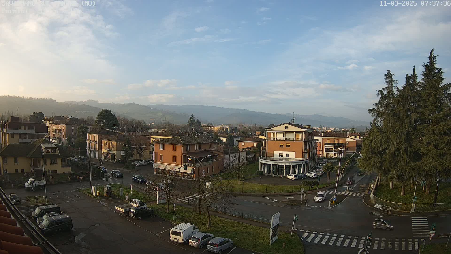 A high-angle, long shot of a town square on a partly cloudy day, surrounded by low buildings and hills in the background.