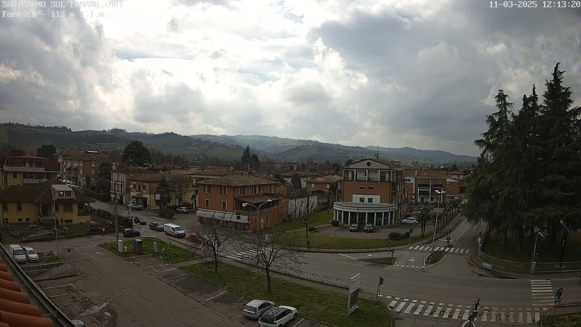A partly cloudy day overlooks a town nestled in a valley, with cars parked in a street-level parking lot and several buildings visible.