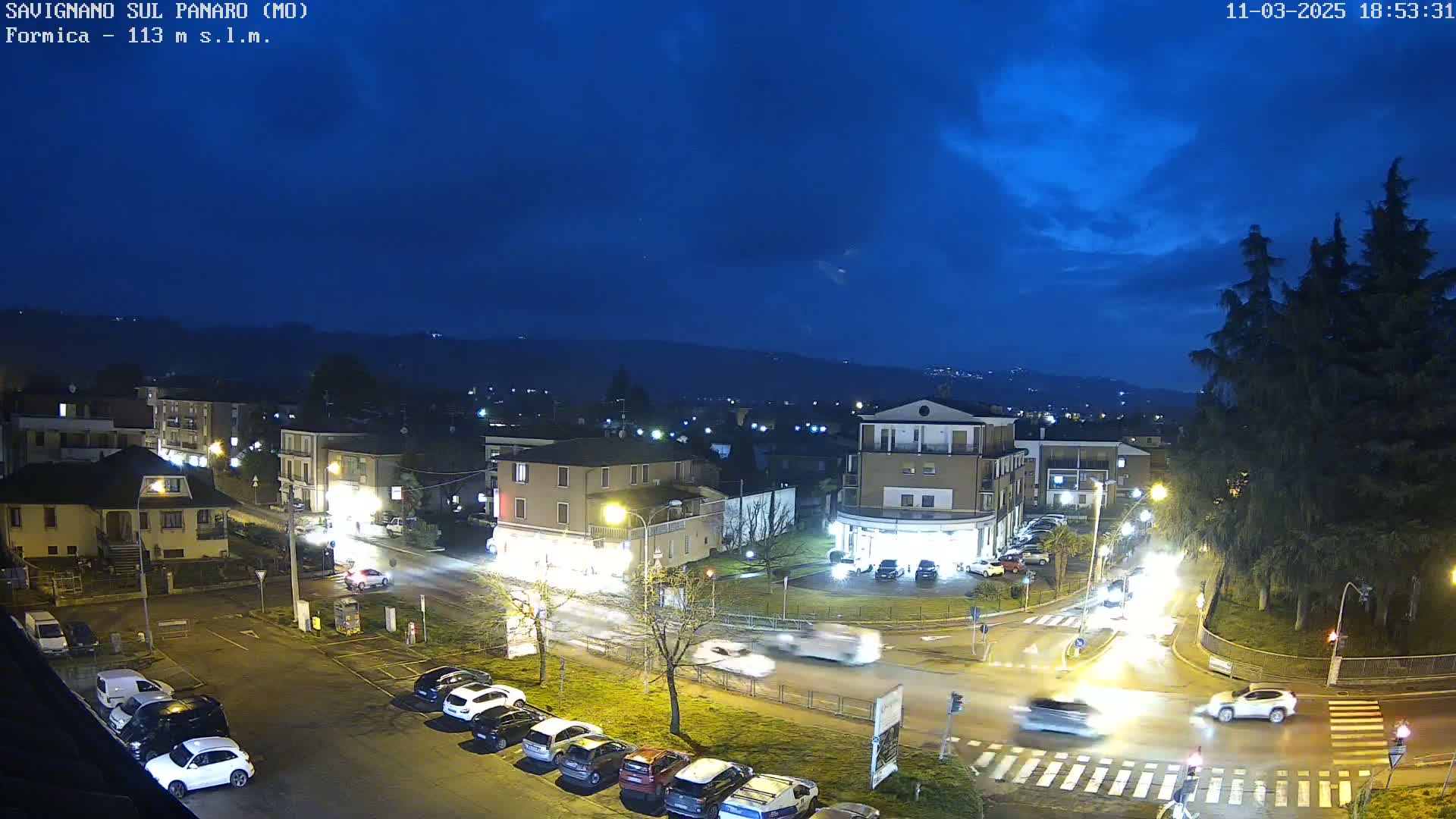 A nighttime aerial view of a town square with cars driving through a lit intersection under a partly cloudy, dark blue sky.