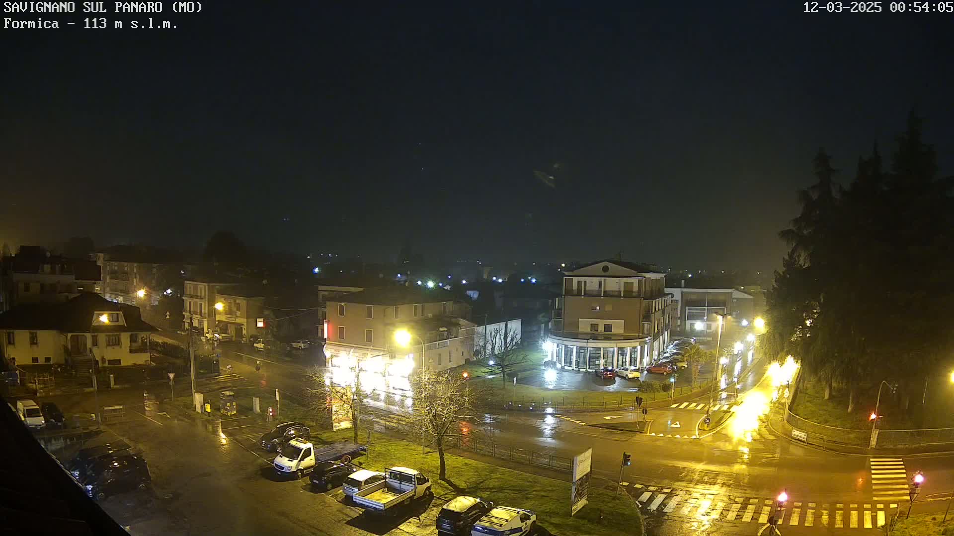 A rain-slicked town square at night, illuminated by streetlights, shows several parked cars and buildings.