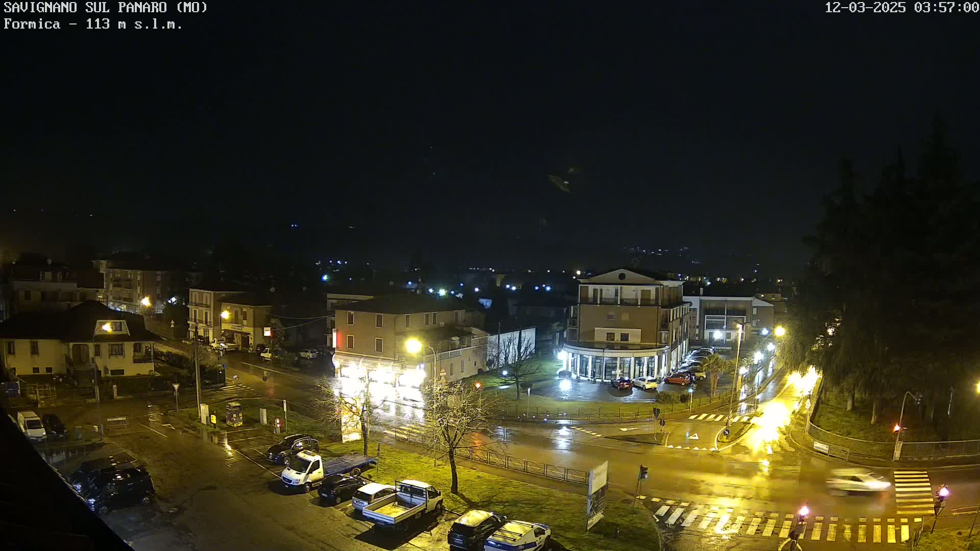A nighttime, rain-slicked town square is illuminated by streetlights, with cars parked and driving along wet roads.