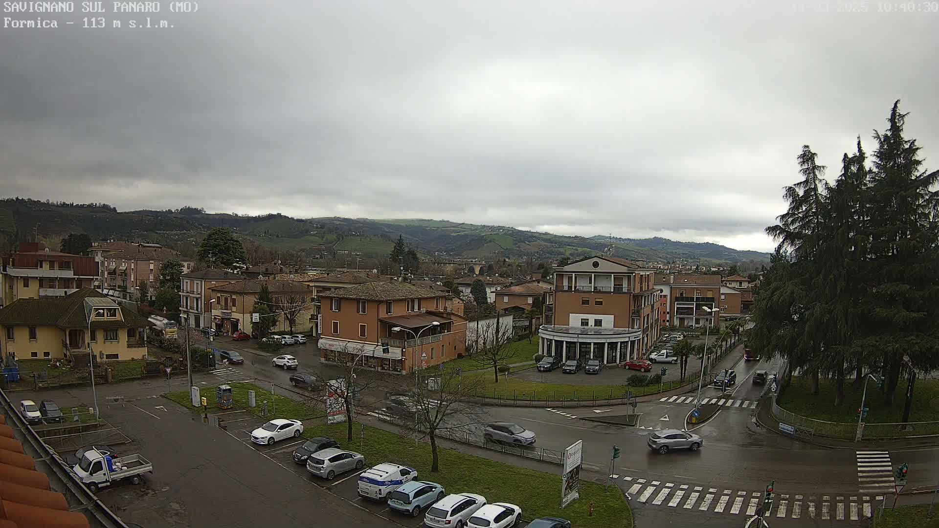 A high-angle view of a town on a cloudy day shows several low-rise buildings, cars parked along streets, and a small roundabout, with rolling hills in the background.