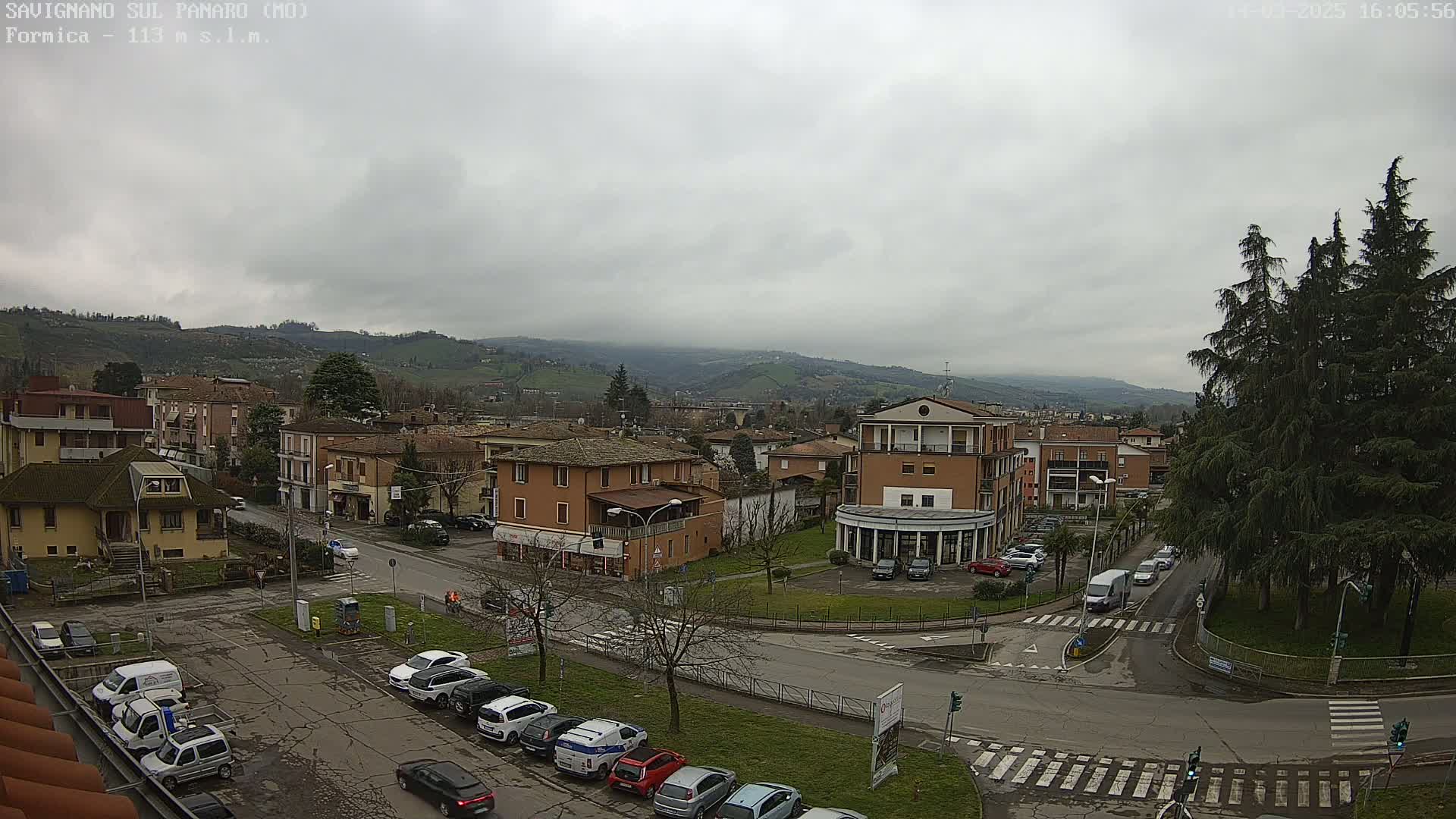 A view of a town situated in a valley under an overcast sky, with cars parked in a lot and driving on roads, surrounded by buildings and trees.
