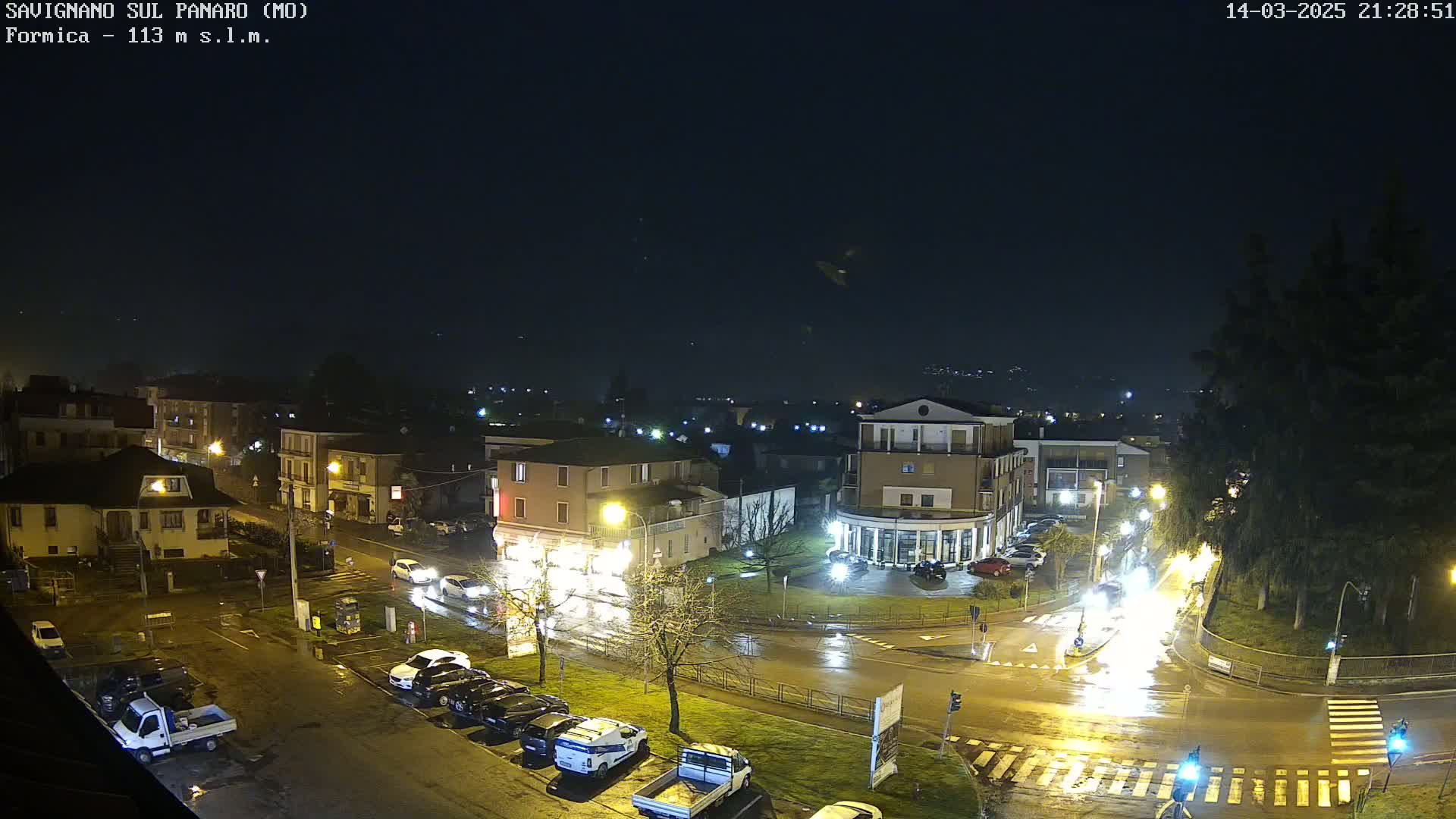 A nighttime view of a town square with wet roads and parked cars under a dark, possibly overcast sky.