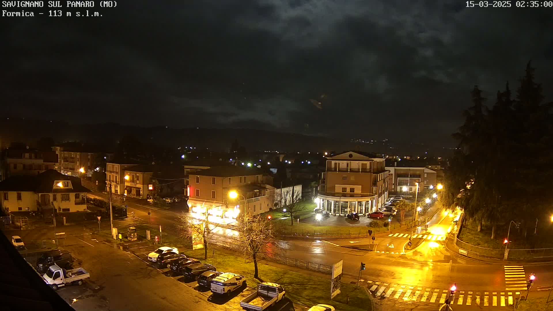 A wet, nighttime view of a town square with buildings, parked cars, and streetlights illuminating the rain-slicked streets.