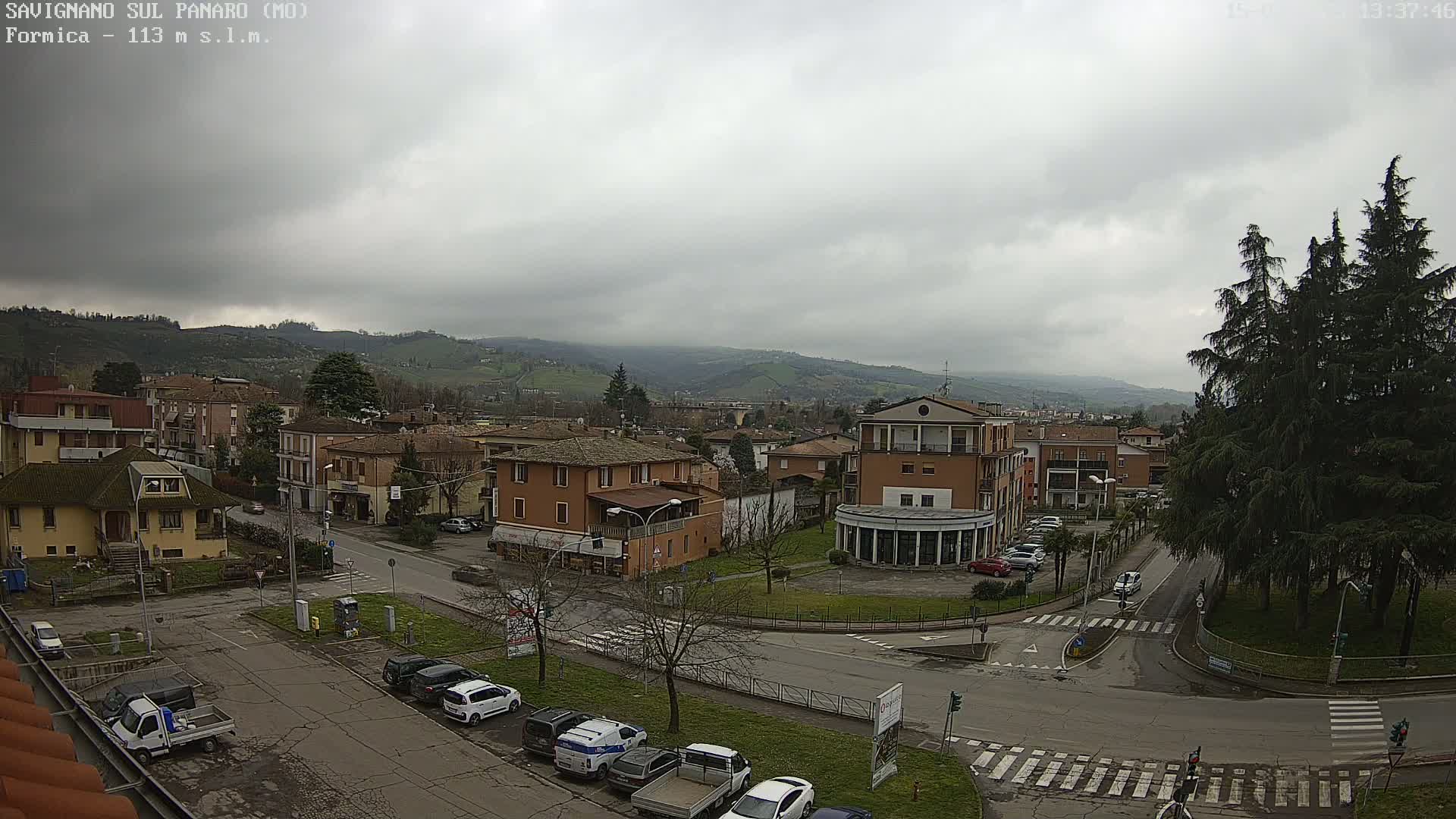 A mostly cloudy day reveals a town nestled in a valley, with cars parked along streets and buildings of varying heights, and evergreen trees in the foreground.